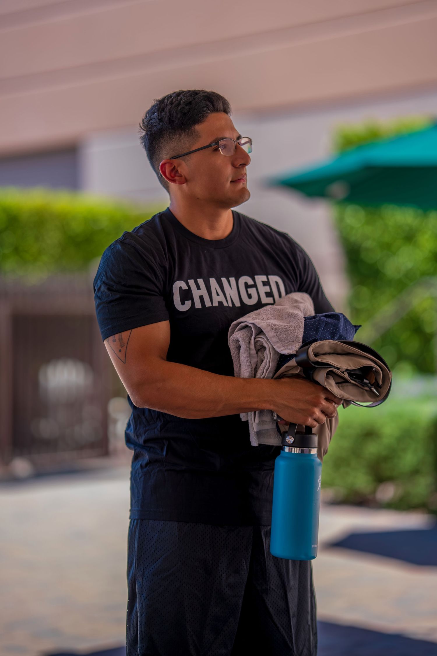 Muscular man in black 'CHANGED' t-shirt holding clothes and blue water bottle outdoors.