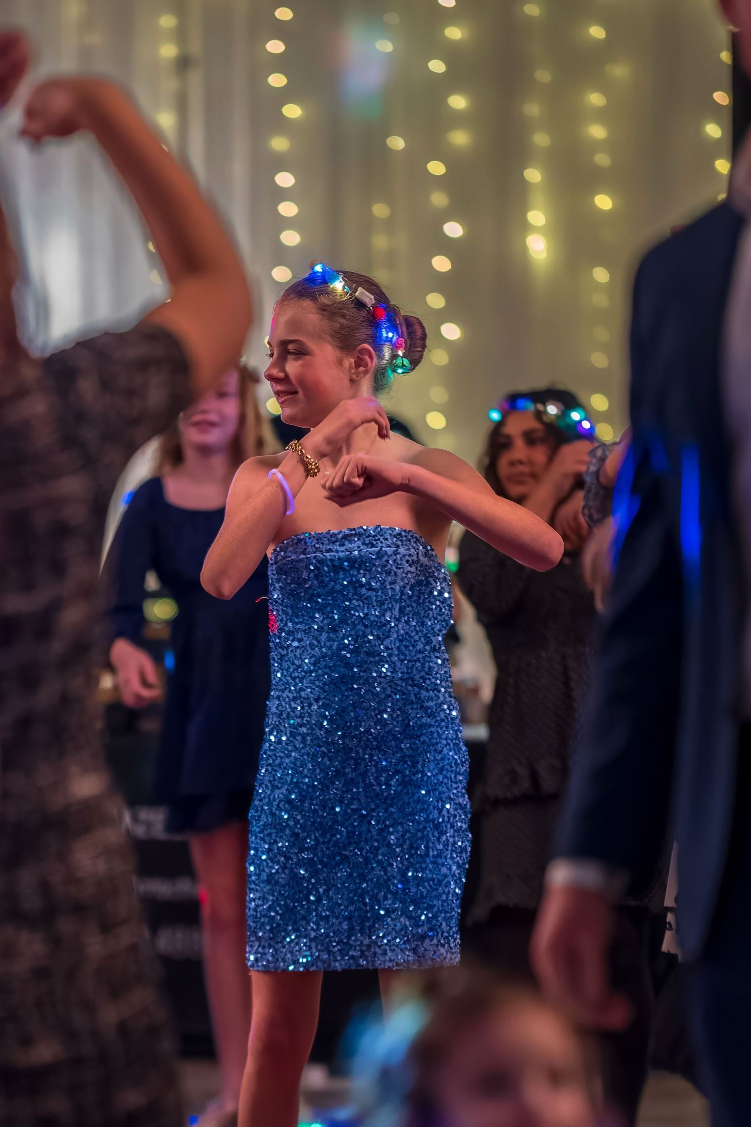 Young girl in blue sequin dress dancing joyfully at a party with string lights in background.