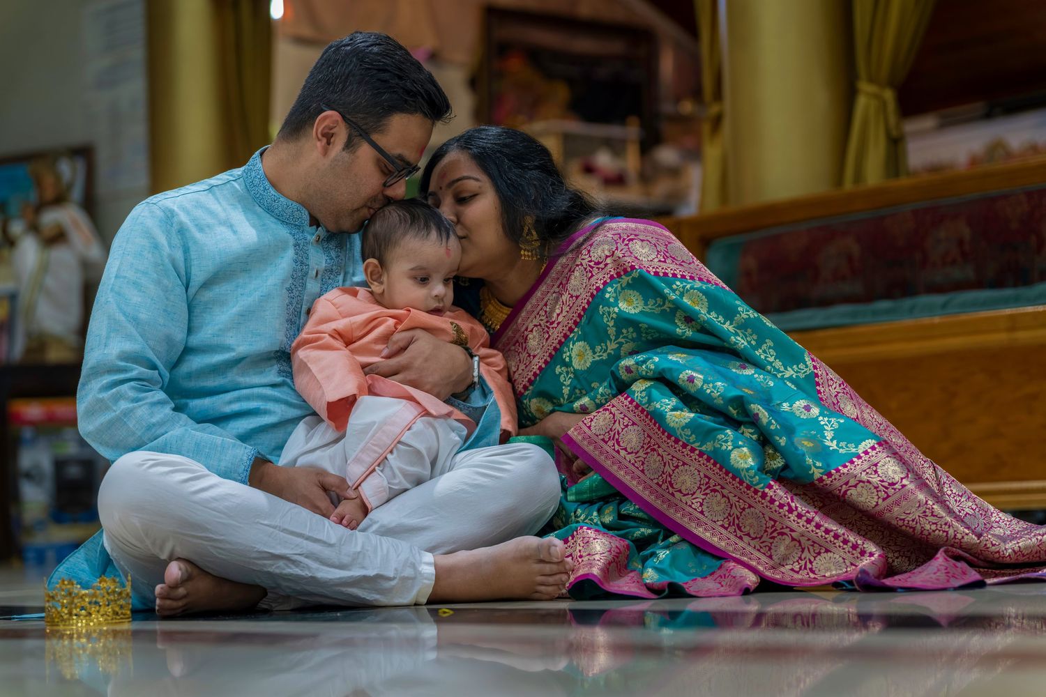Indian couple in traditional attire sitting on floor sharing tender moment with baby in pink outfit.