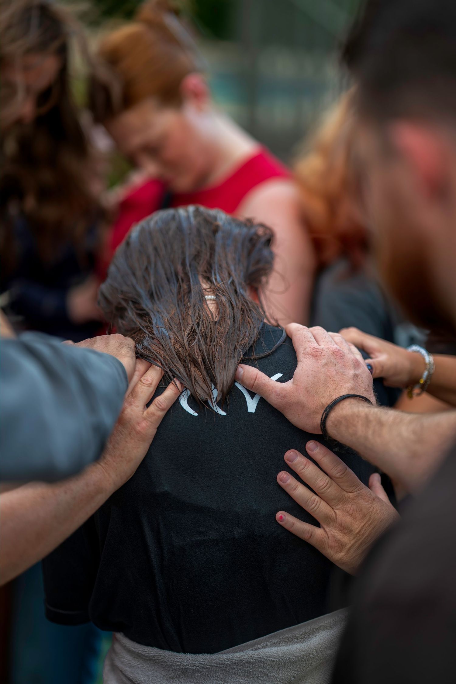 Group of people placing hands on person's back in prayer circle outdoors.