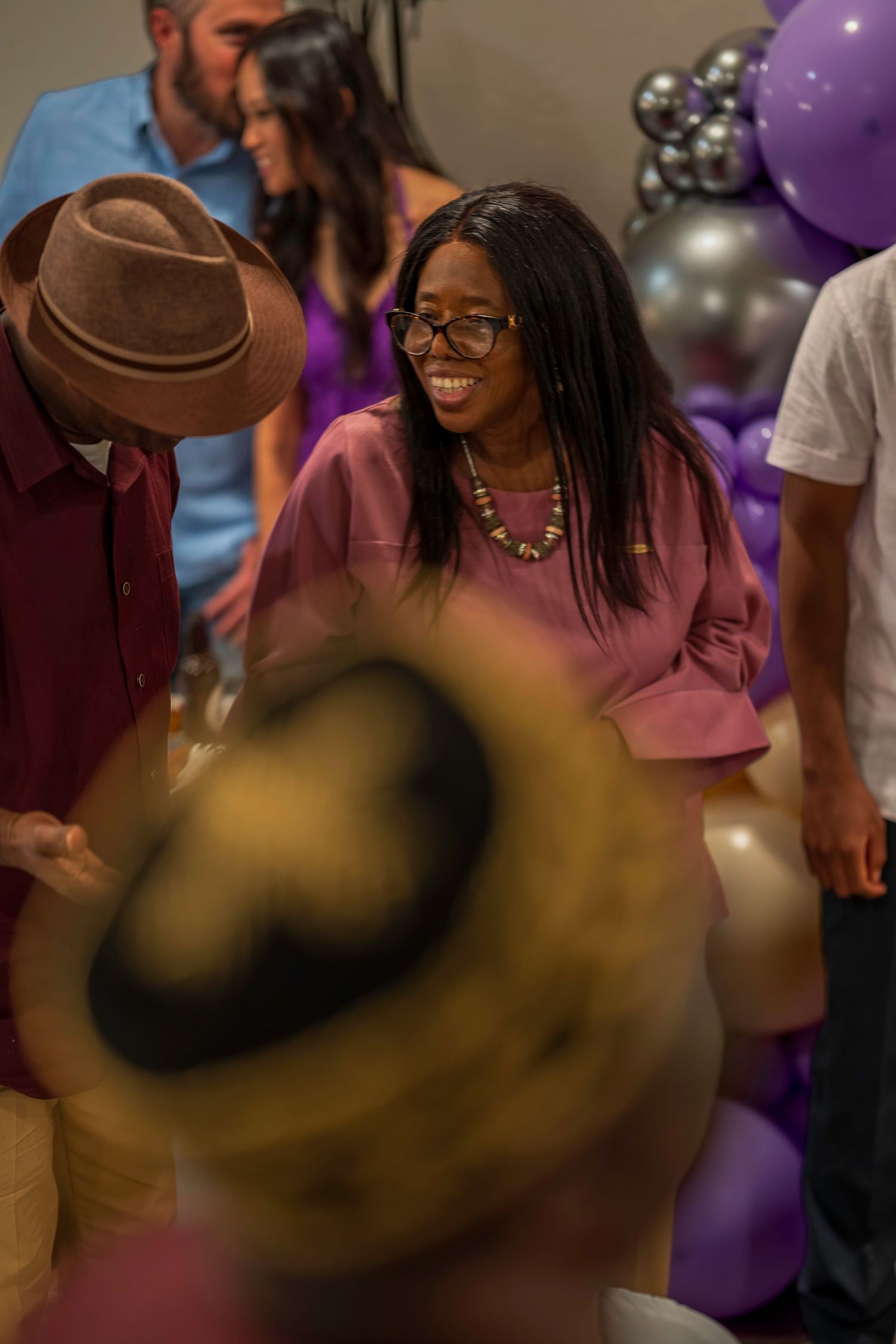 Smiling Black woman in purple top and glasses at a festive party with purple and silver balloons.