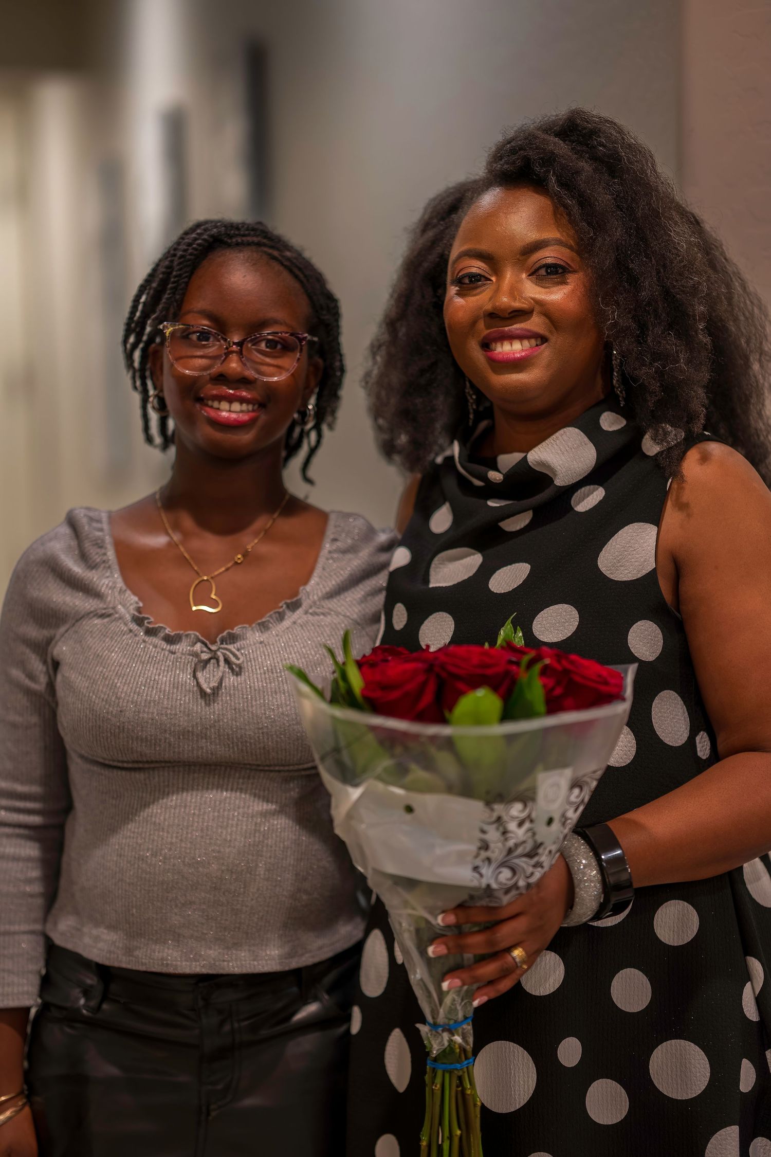 Two smiling women pose together, one holding a bouquet of red roses at a celebration.