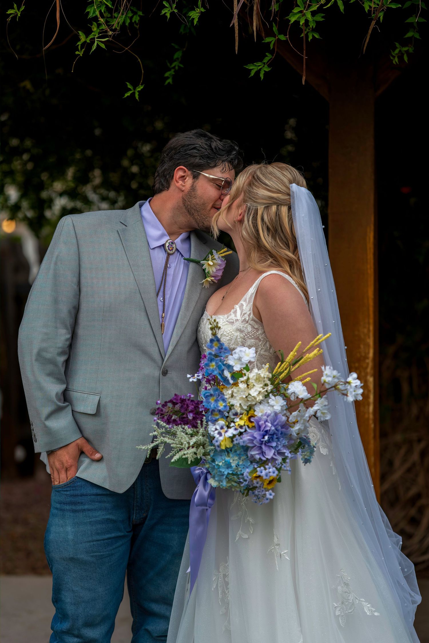 A bride and groom sharing a kiss under a vine-covered arbor, bride holding a colorful wildflower bouquet.