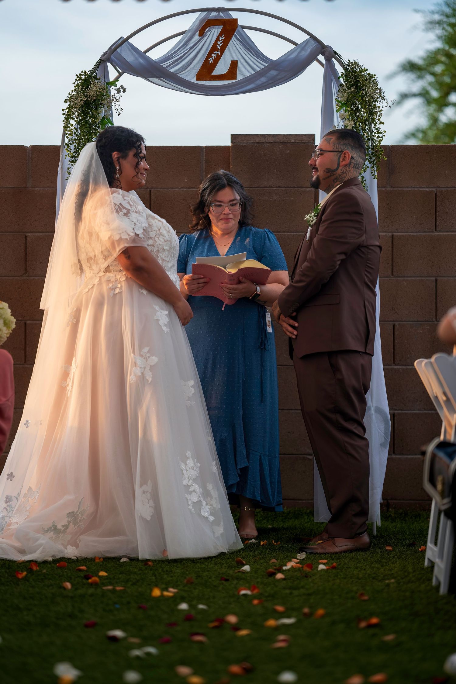 Bride and groom exchange vows under floral arch with officiant at outdoor backyard wedding ceremony.