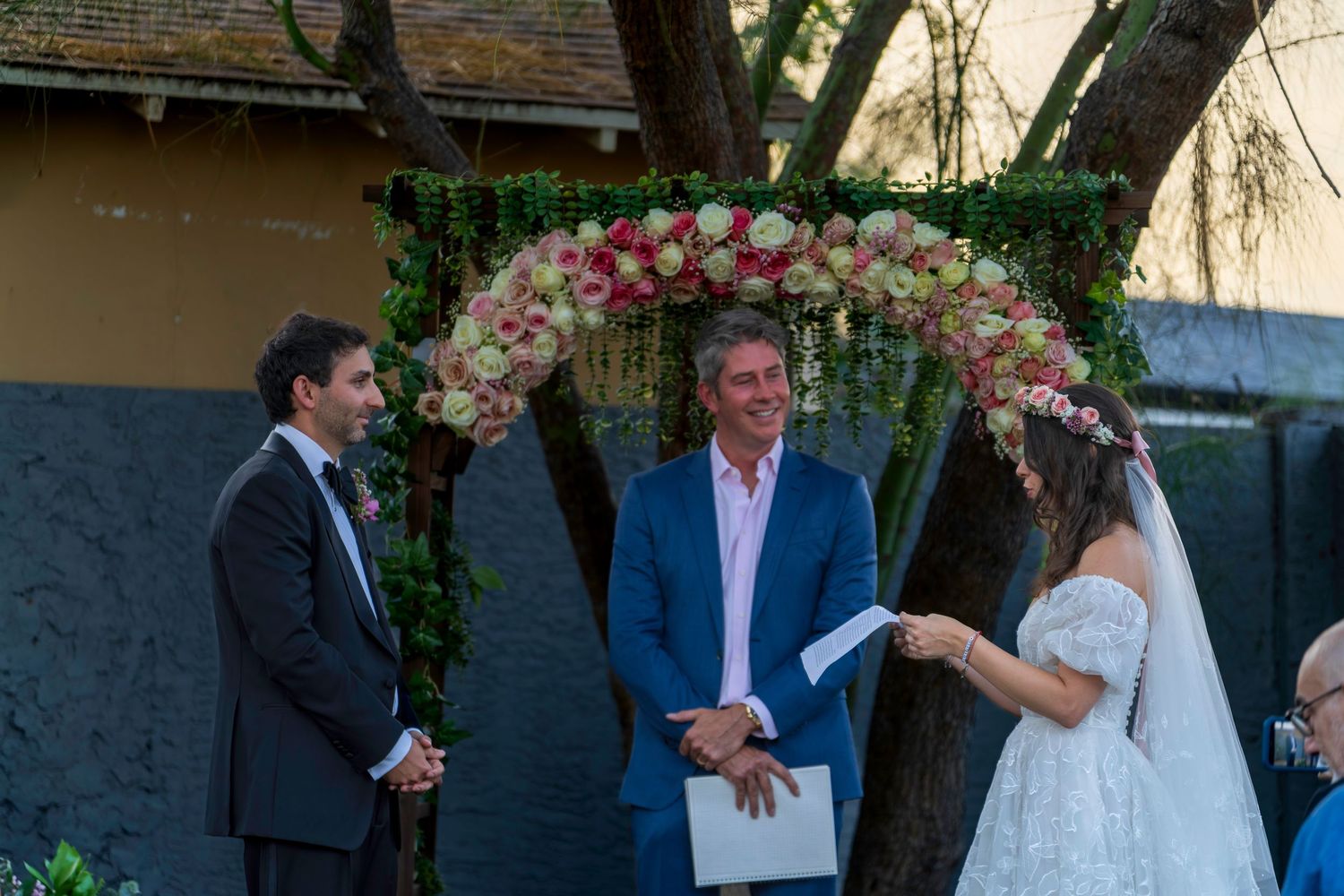 Couple exchanging vows outdoors under floral arch with officiant at elegant wedding ceremony.