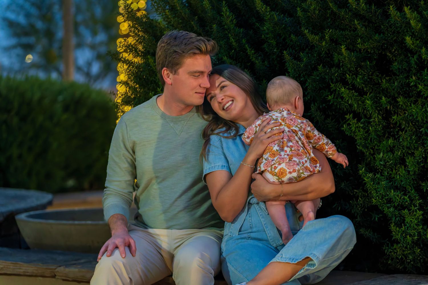 Happy couple with baby sitting together near illuminated garden at dusk.
