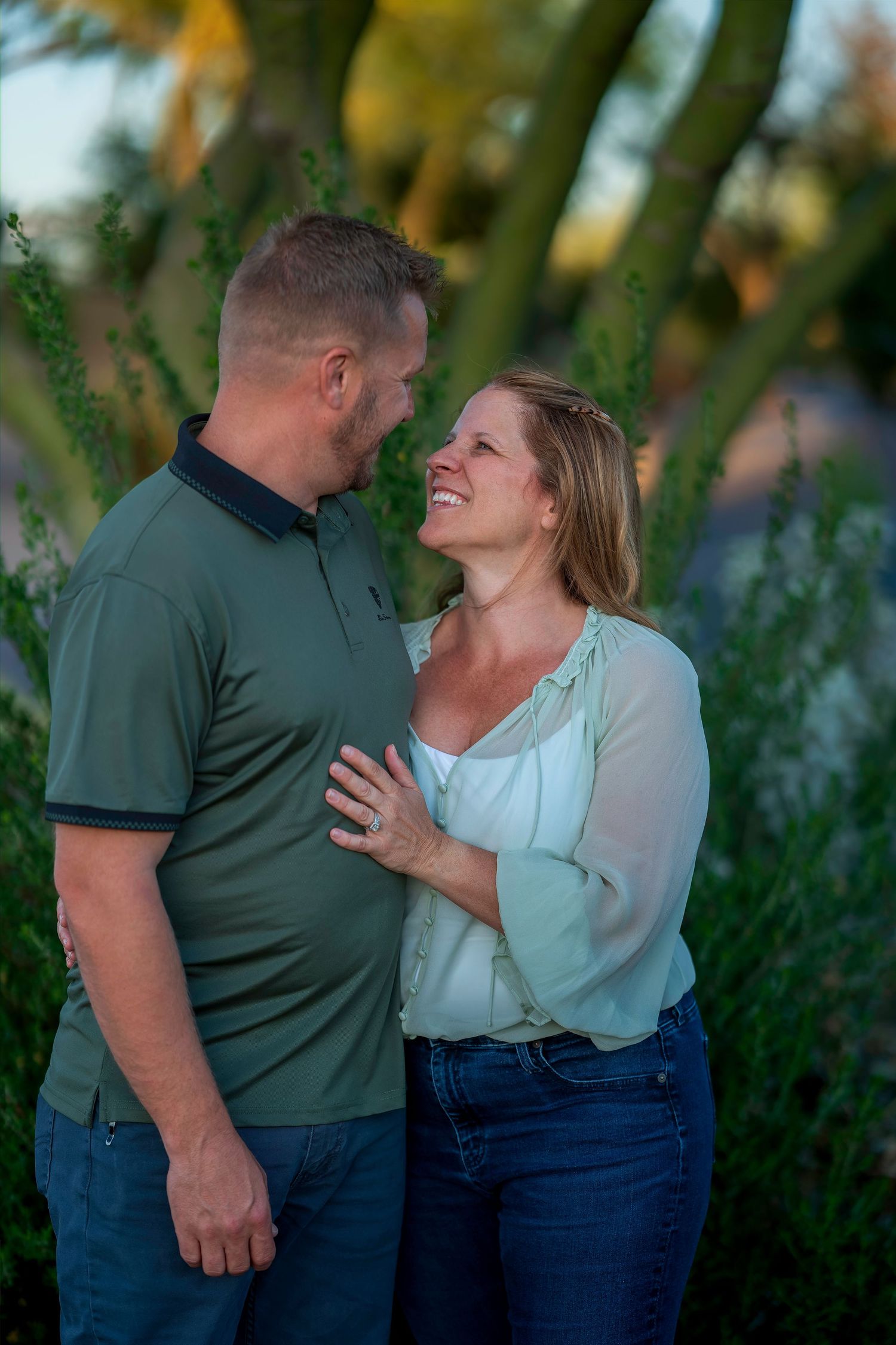 A smiling couple in casual attire shares a tender moment outdoors, surrounded by lush green foliage at golden hour.