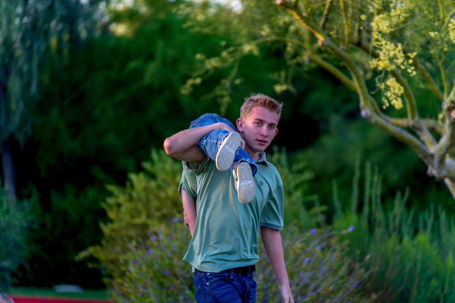 Young teenage boy in a green polo shirt carrying shoes over his shoulder in a lush garden setting.
