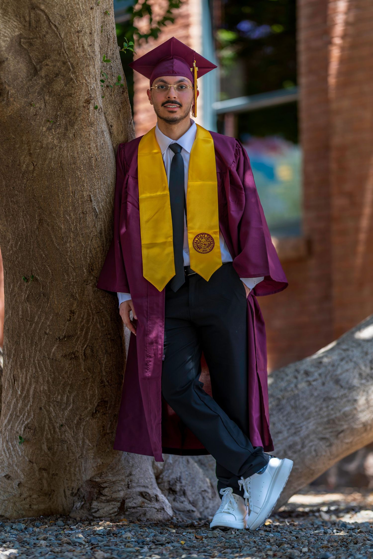 Young man in maroon graduation gown and gold stole leaning against tree outdoors.