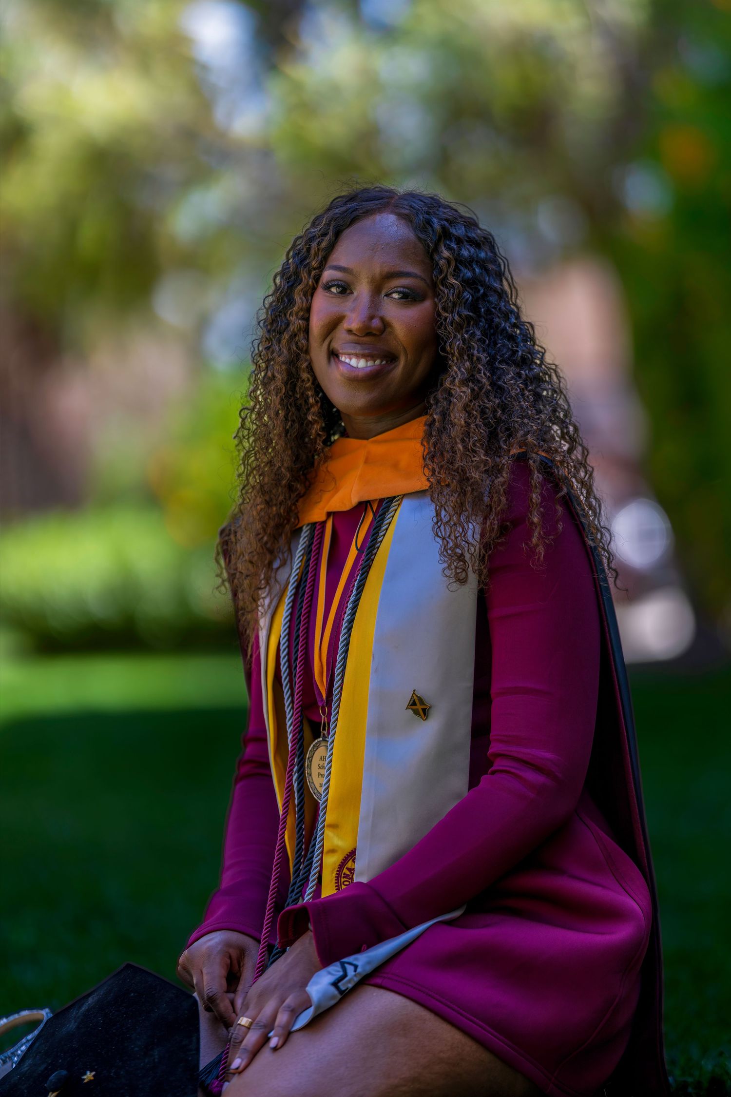 Smiling ASU graduate in maroon doctoral gown with gold and white stole seated on green campus lawn.