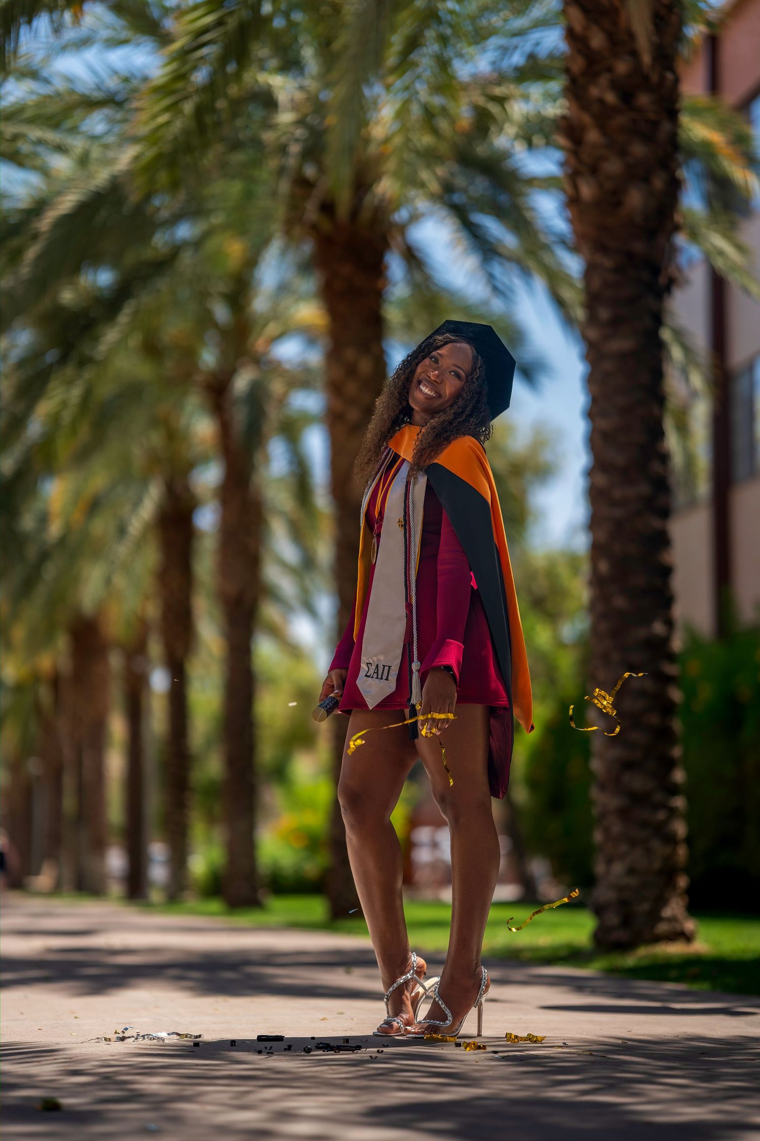 A smiling graduate poses in maroon dress and graduation regalia among tall palm trees on a sunny campus path.