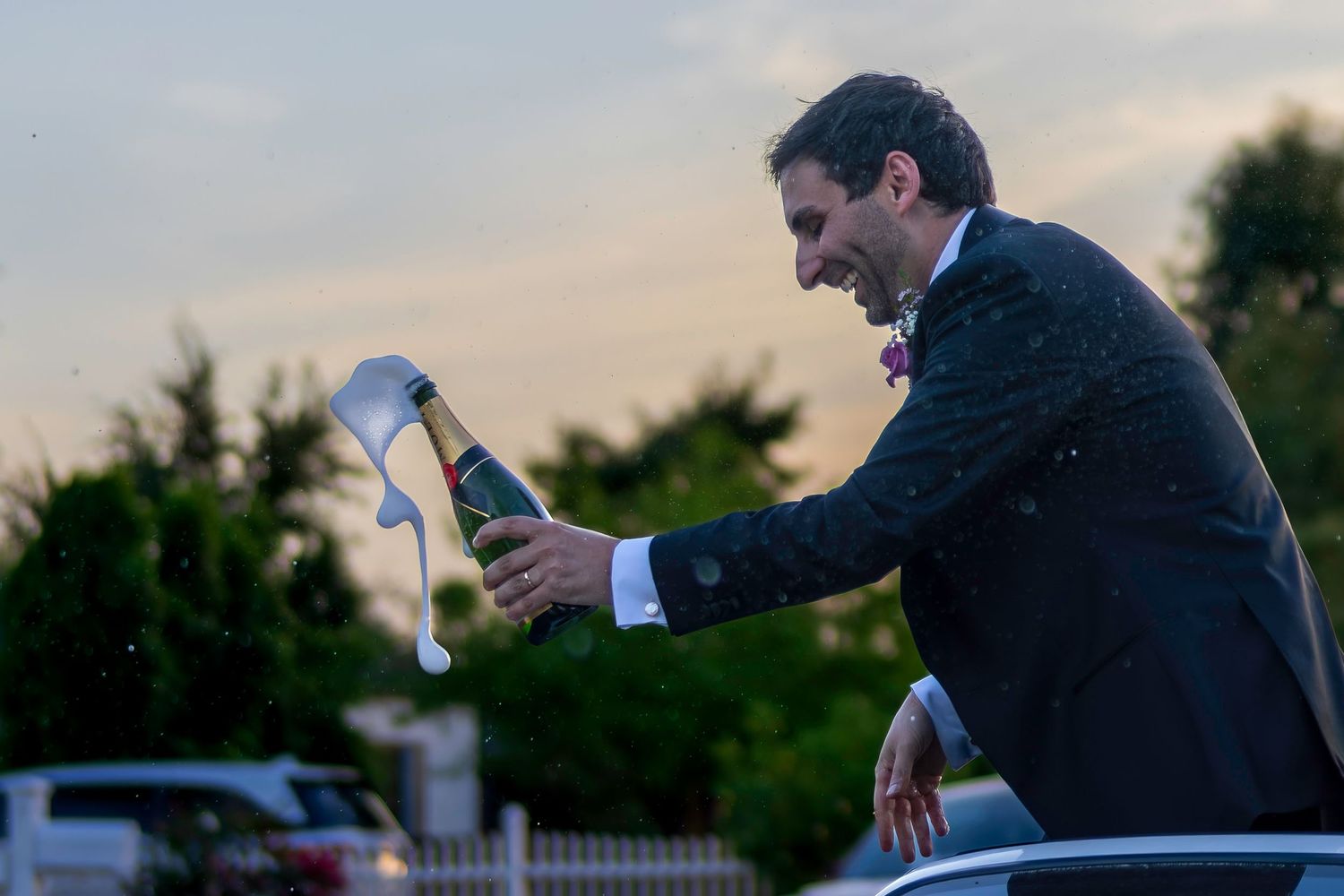 Groom in tuxedo celebrating with champagne bottle popping outdoors at dusk during wedding reception.