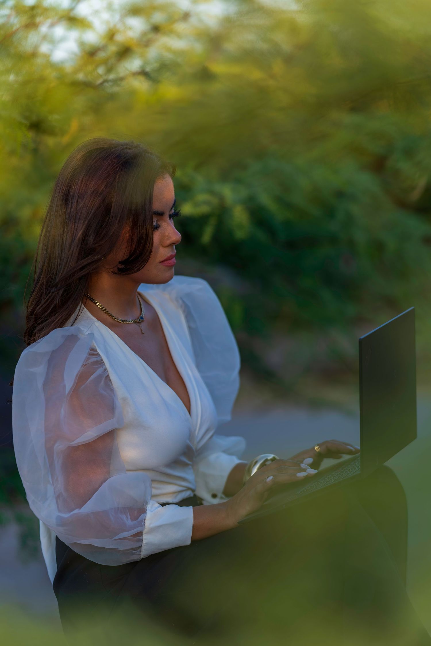 Woman in white sheer blouse working on laptop outdoors surrounded by lush green foliage in natural light.
