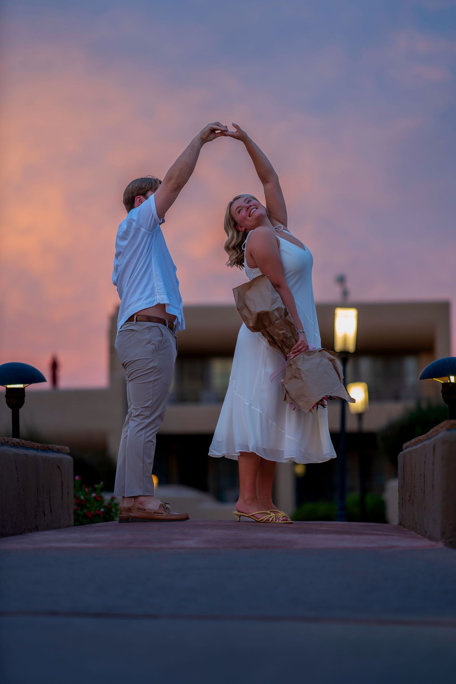 Couple dancing at sunset, woman in white dress twirling under man's hand, colorful pink sky backdrop.