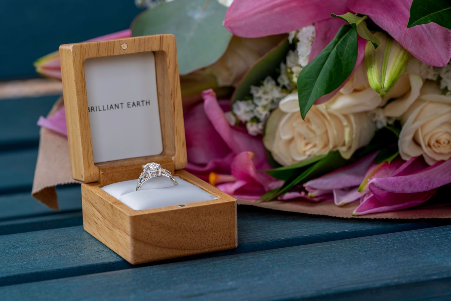 Brilliant Earth diamond engagement ring in wooden box beside a bouquet of pink and white flowers.