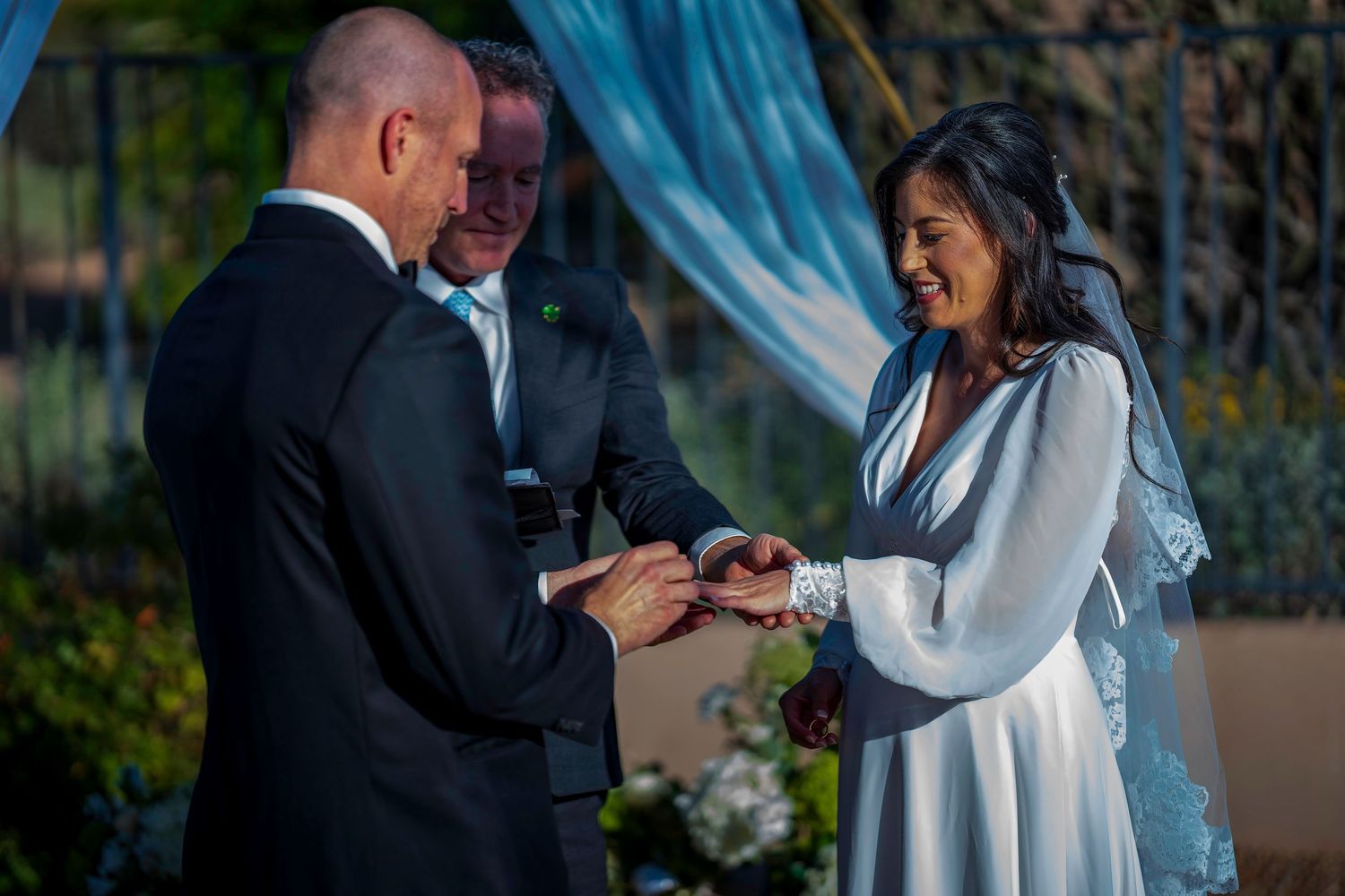 Groom places wedding ring on smiling bride's finger during outdoor ceremony under blue sky.