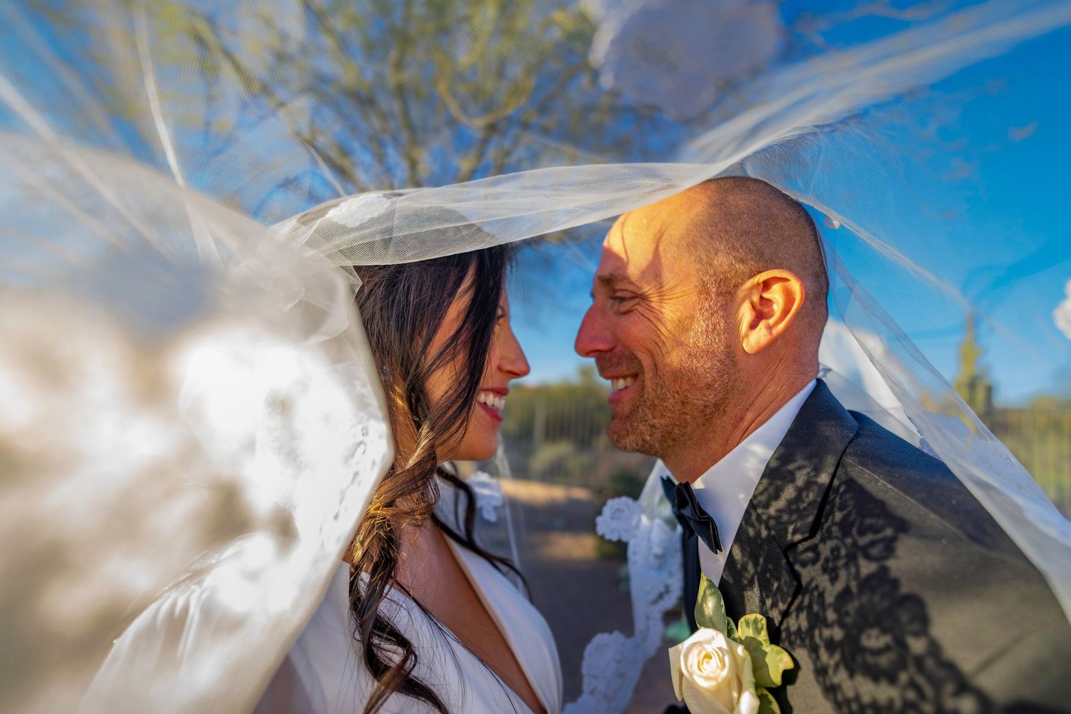 Bride and groom laugh joyfully under flowing white veil against bright blue sky backdrop.