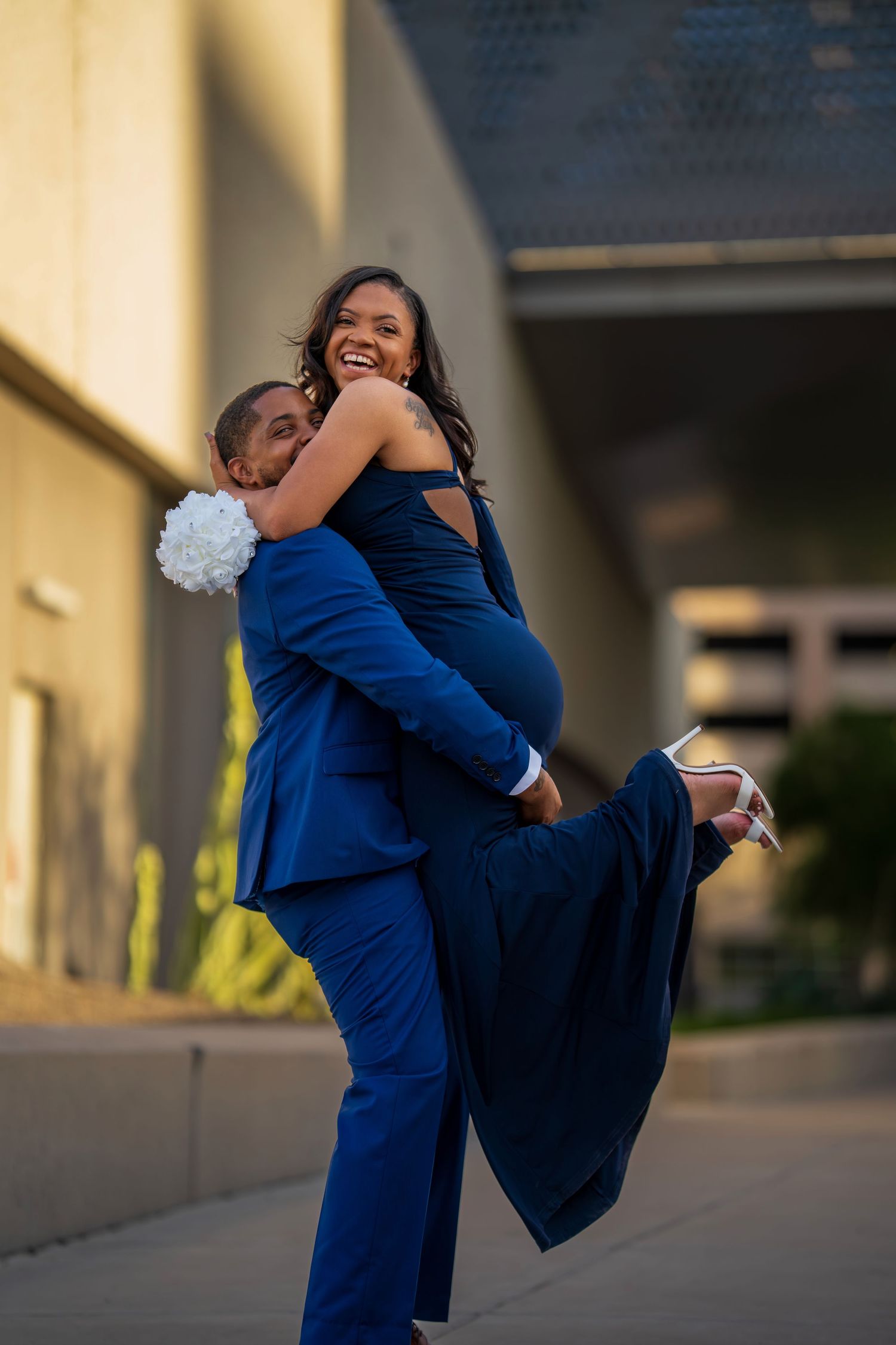 A joyful couple in matching blue outfits, man lifting woman who holds white bouquet, during golden hour outdoors.