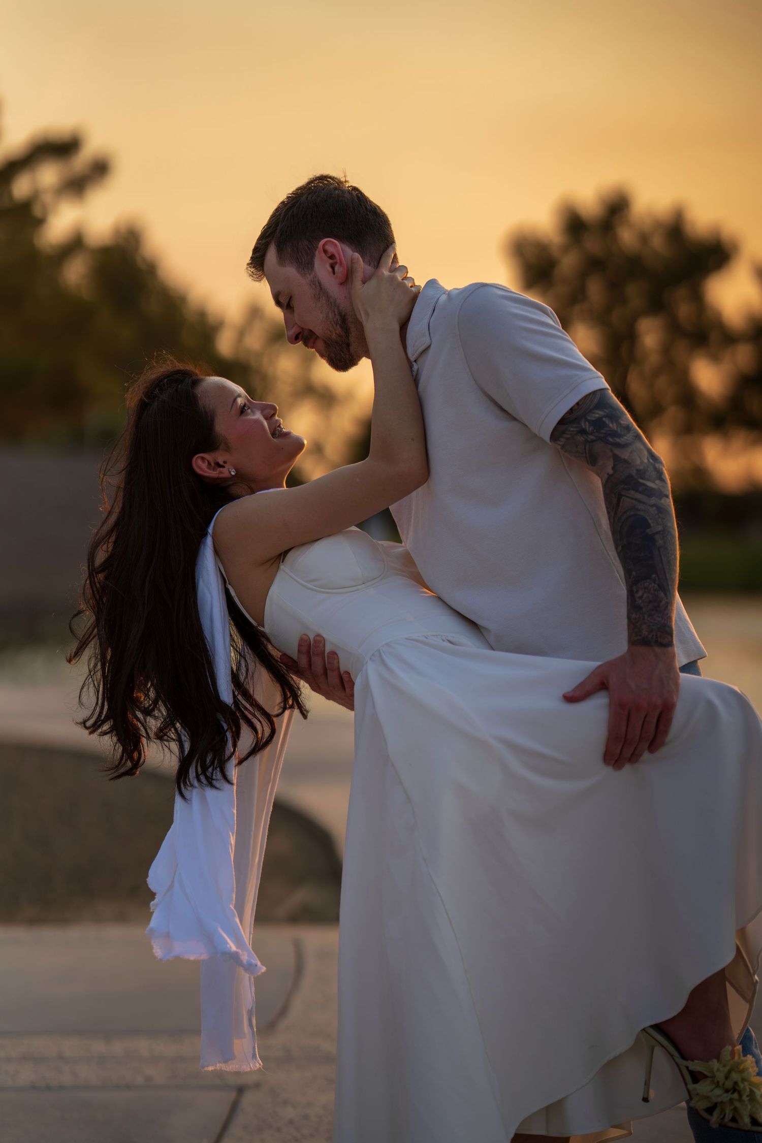 Romantic couple sharing a kiss at golden sunset, woman in white dress with flowing hair, man with tattoos.