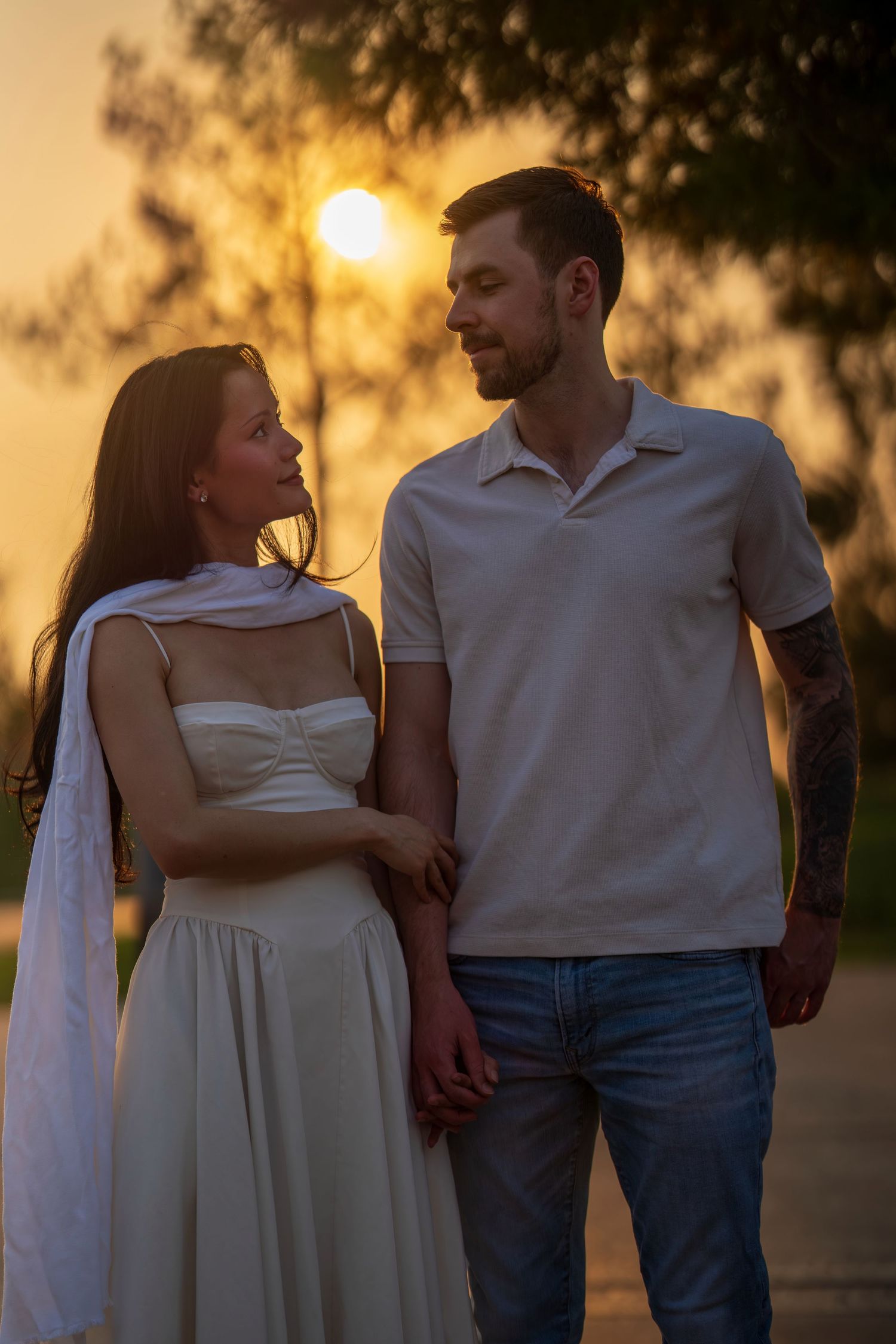 Couple holding hands at golden sunset in park, woman in white dress, man in polo shirt and jeans.