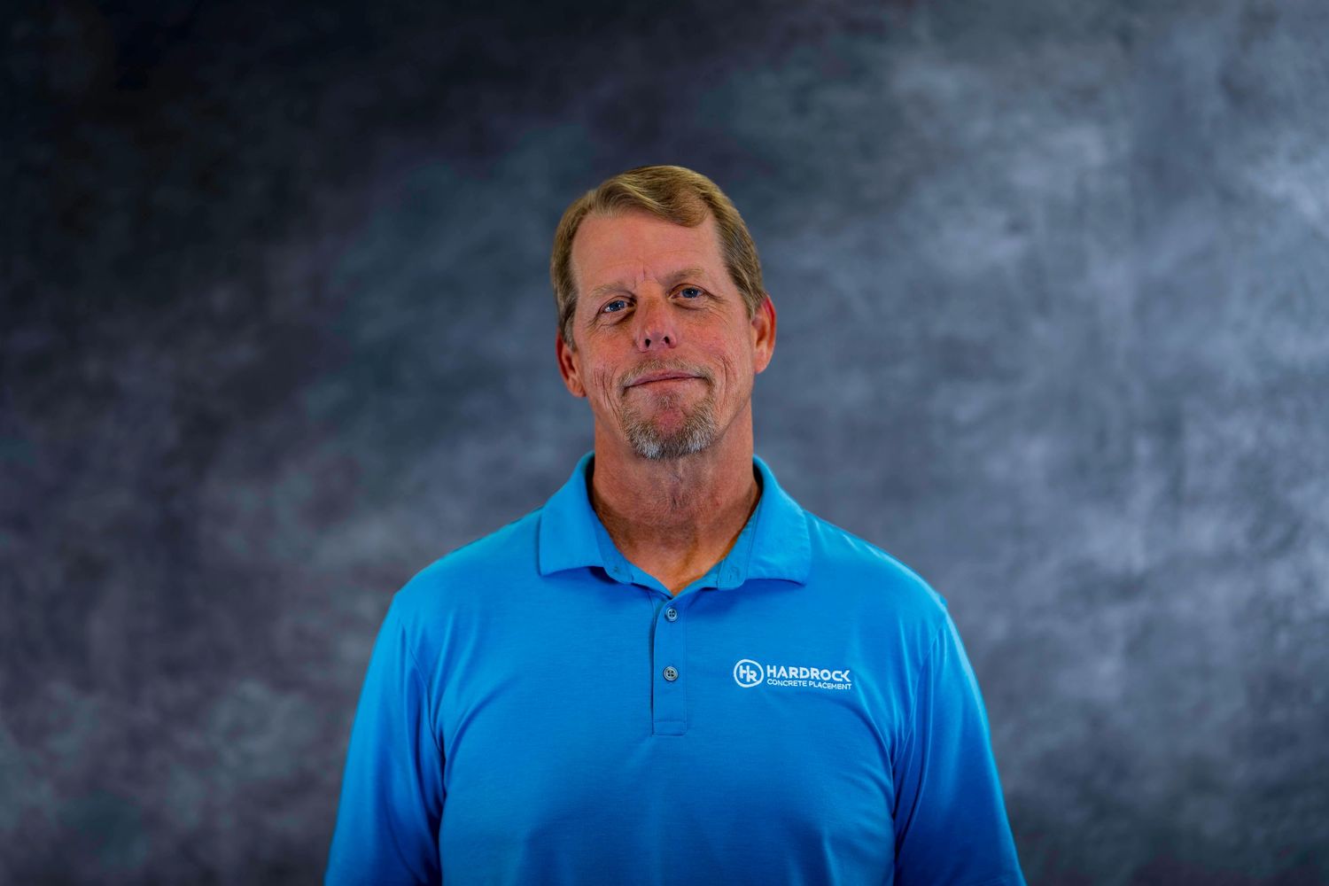 Staff member wearing light blue polo shirt with company logo against gray background.