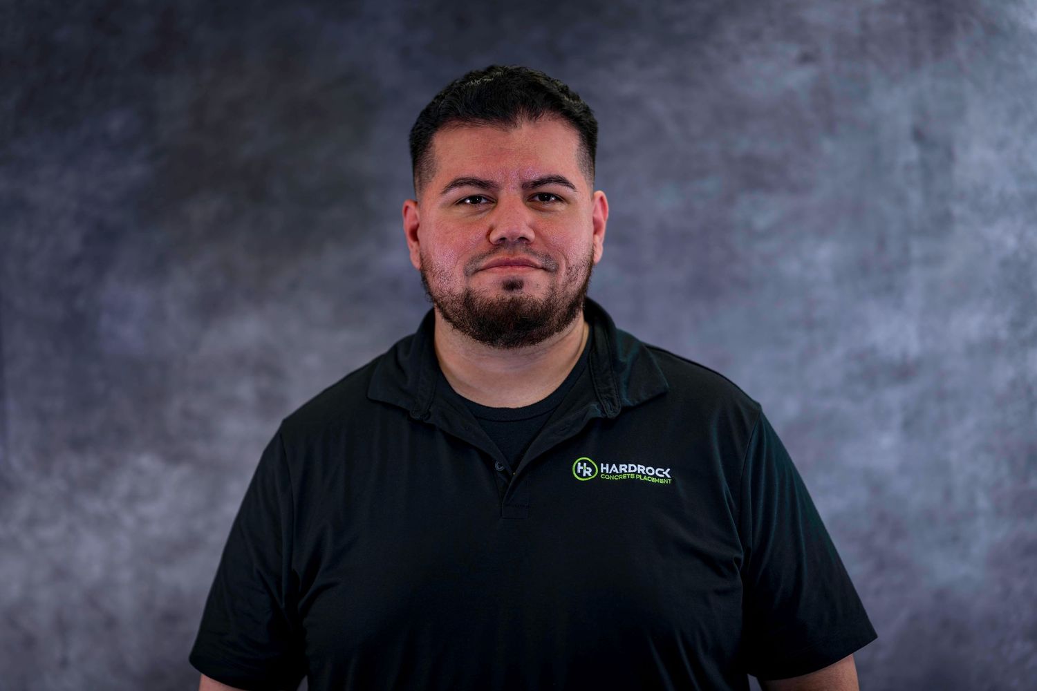 Corporate headshot of person wearing black polo with company branding against gray backdrop.