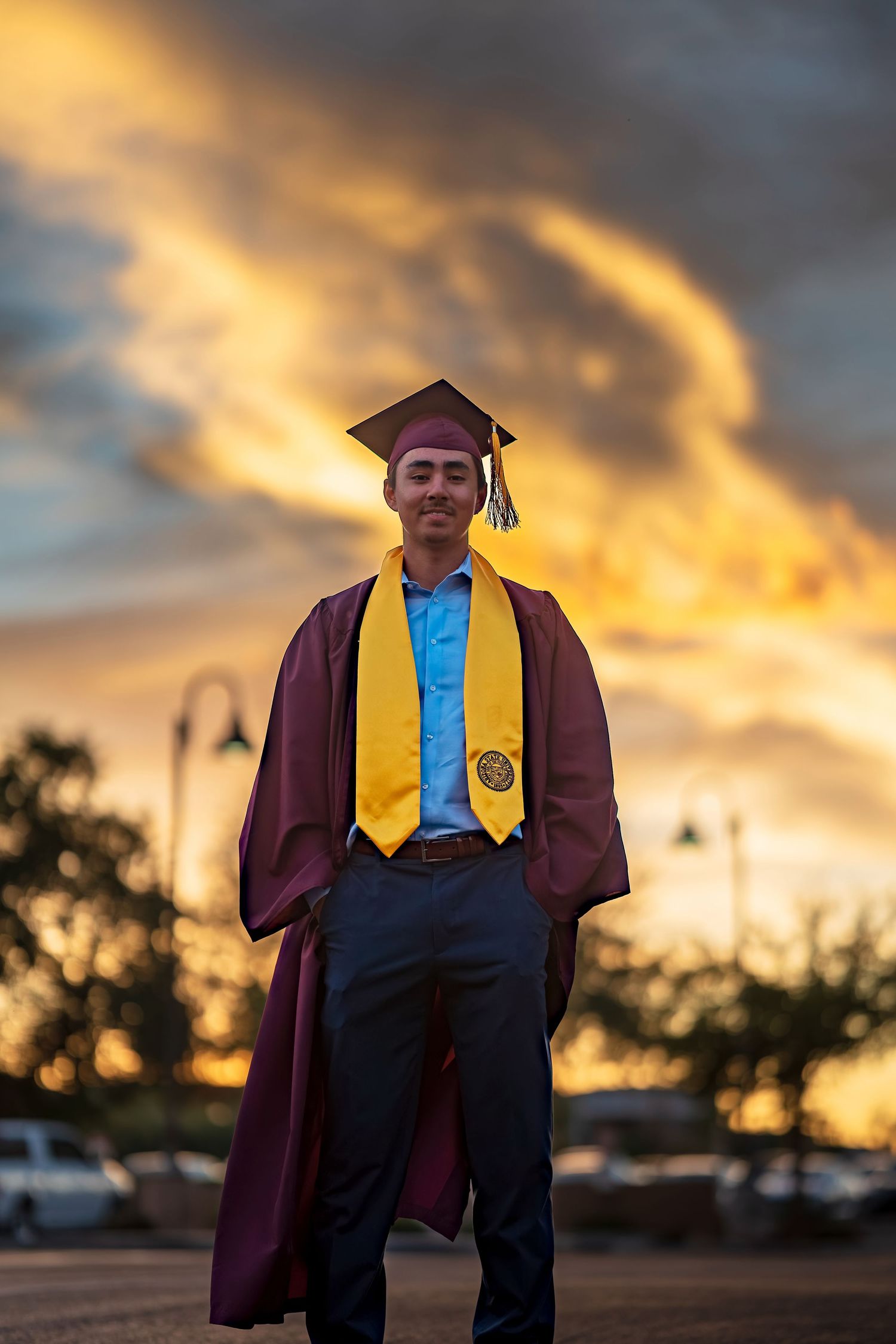 Graduation portrait with maroon cap and gown against golden evening sky.