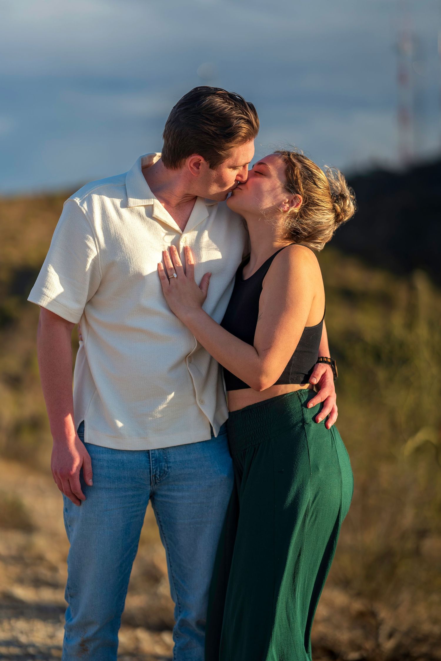 Couple sharing a romantic kiss outdoors at golden hour with hills in the background.
