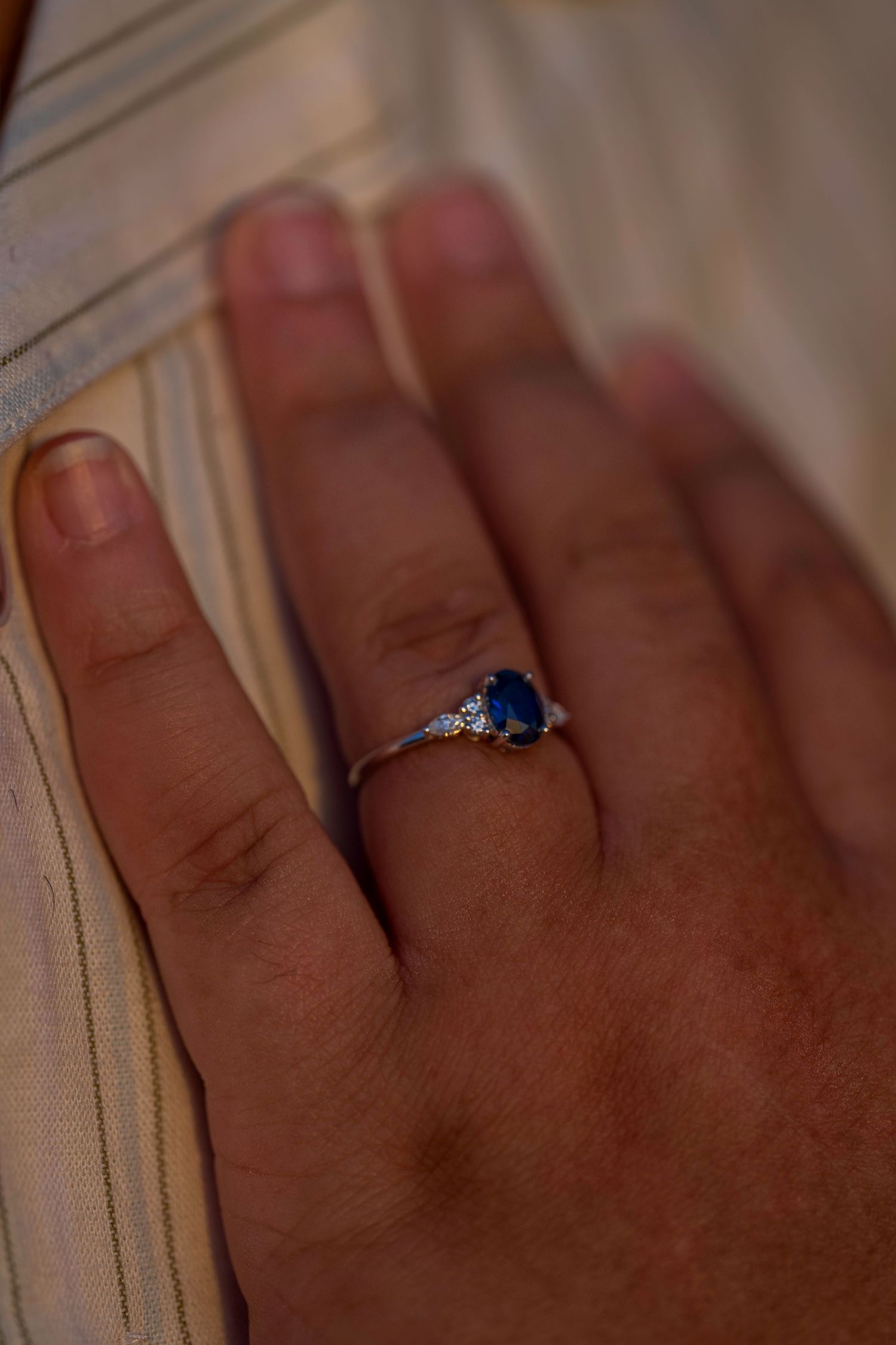 Close-up of hand wearing a blue sapphire and diamond engagement ring against a white fabric background.