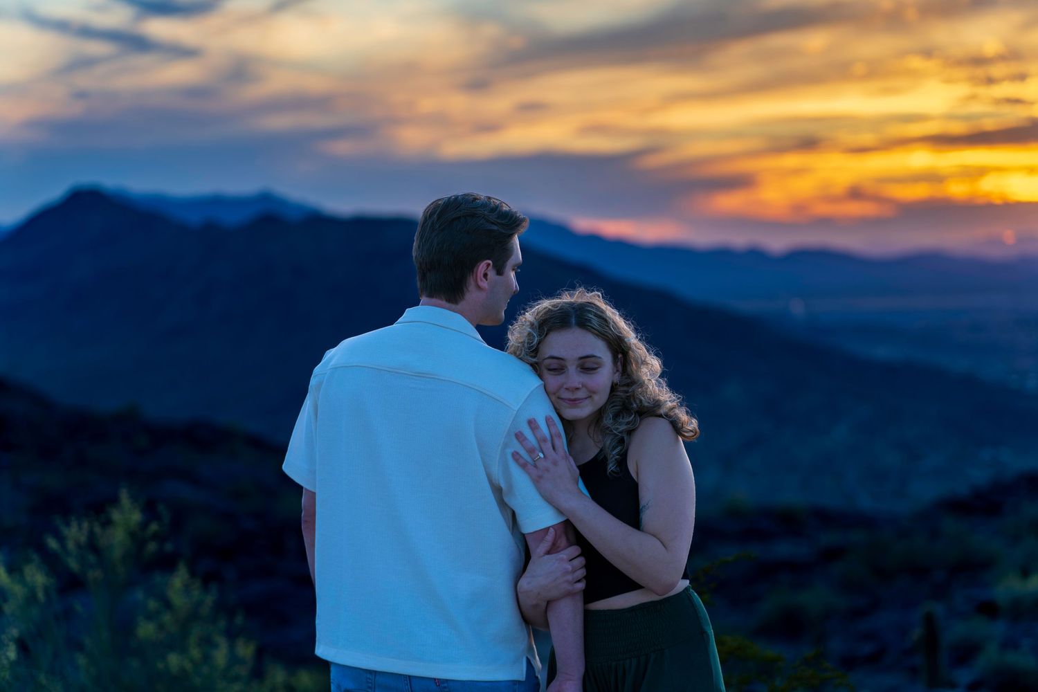 Couple embracing while gazing at a dramatic colorful sunset over mountain landscape.