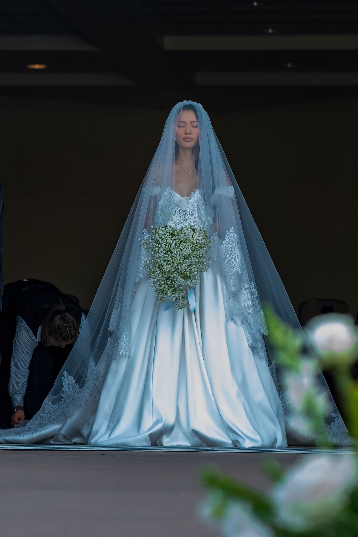 A bride in an elegant white satin wedding gown and long veil holds a baby's breath bouquet during her ceremony.
