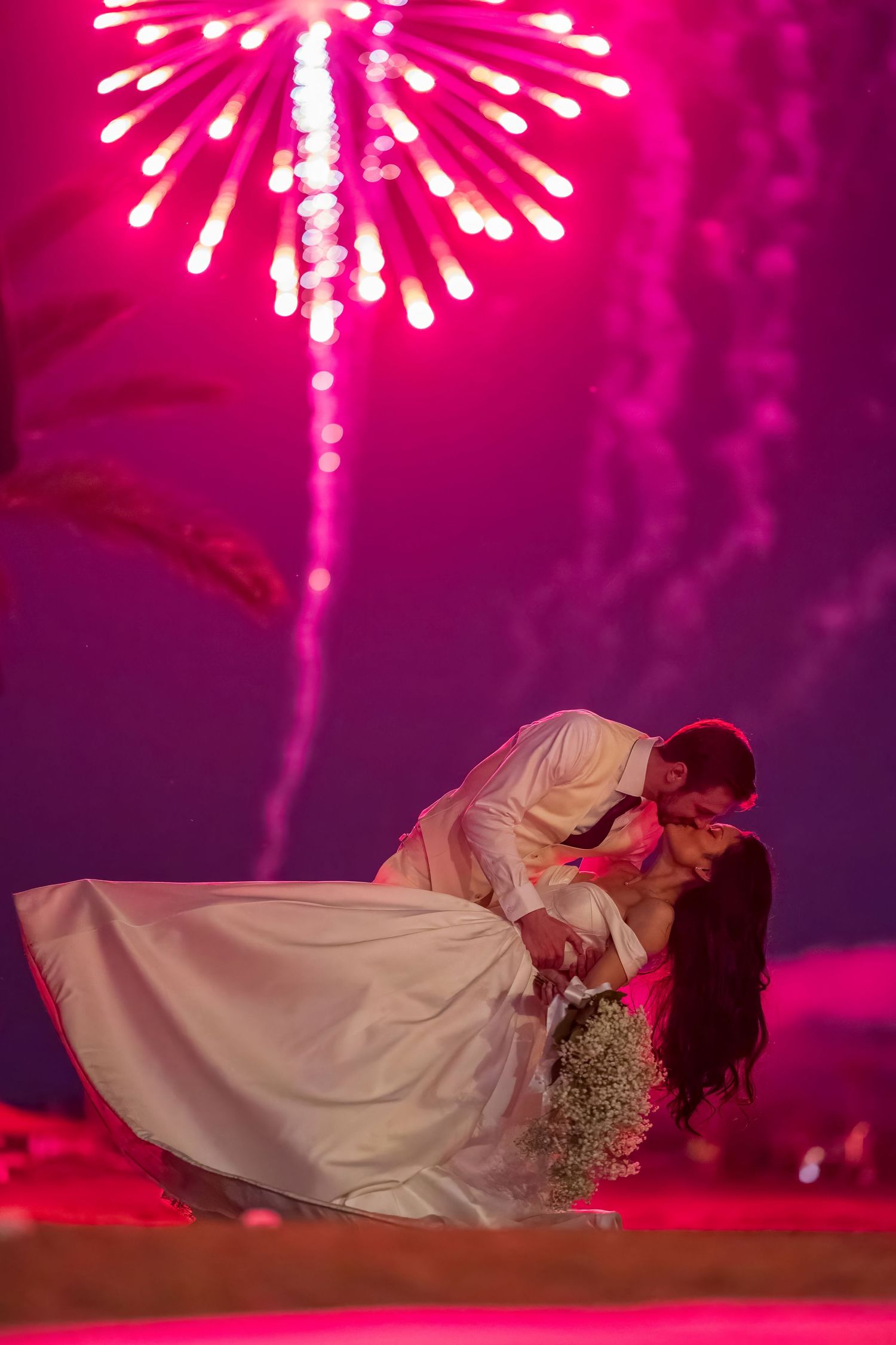 Wedding couple shares intimate moment beneath vibrant pink fireworks in the night sky.