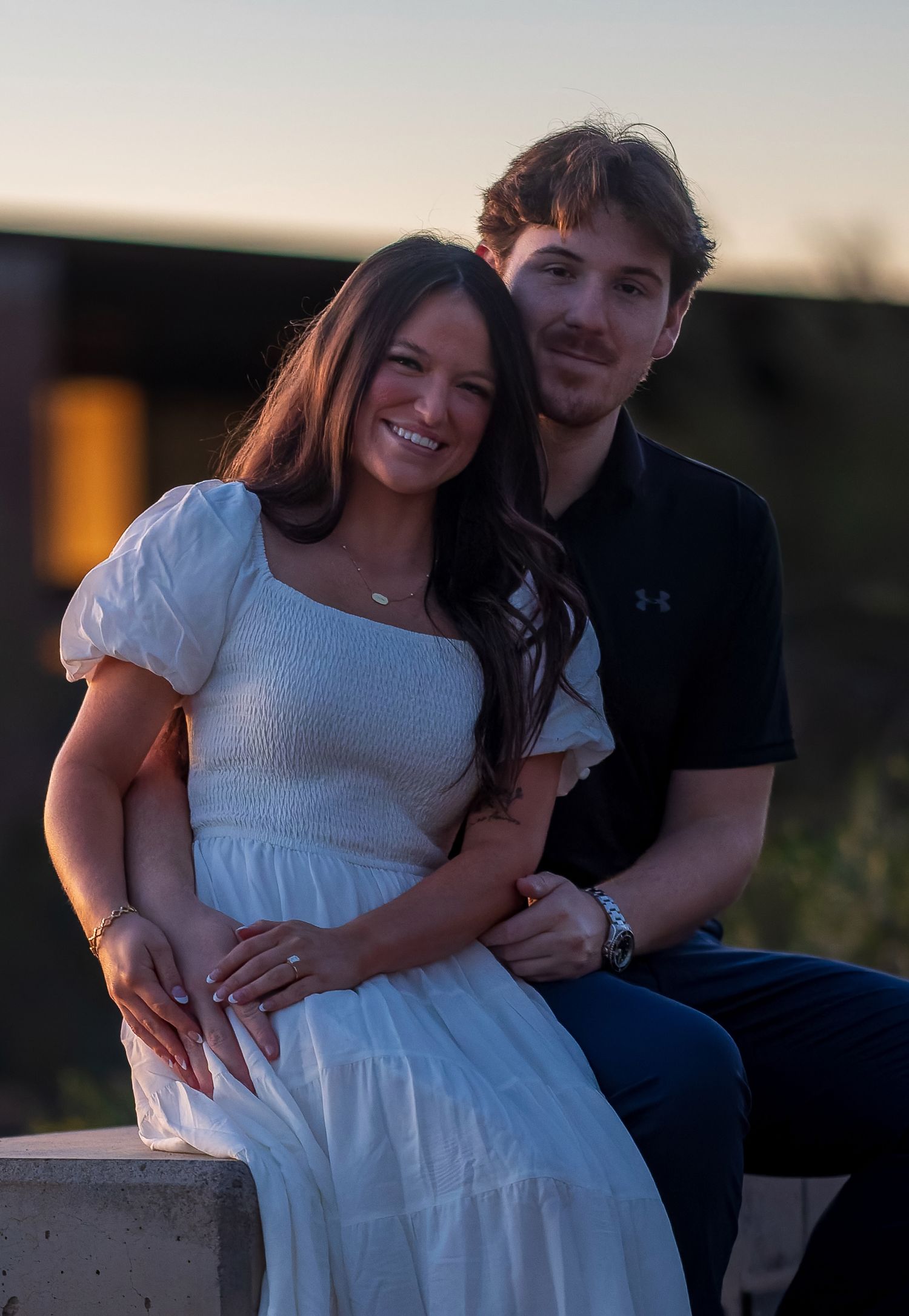 Romantic couple embraces during sunset photoshoot, with woman in white dress and man in black shirt.