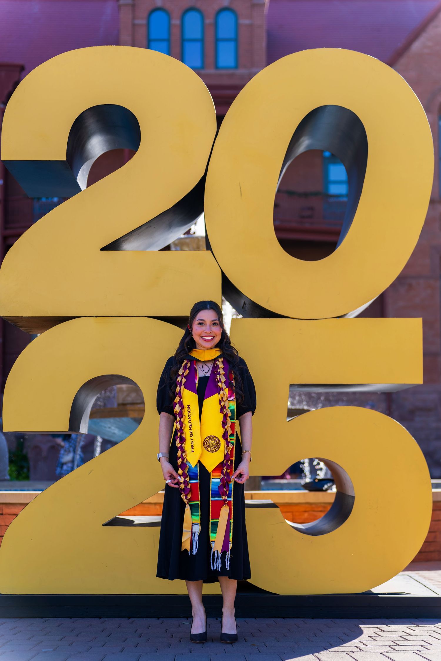A graduate poses in front of large yellow 2025 numbers on a building.