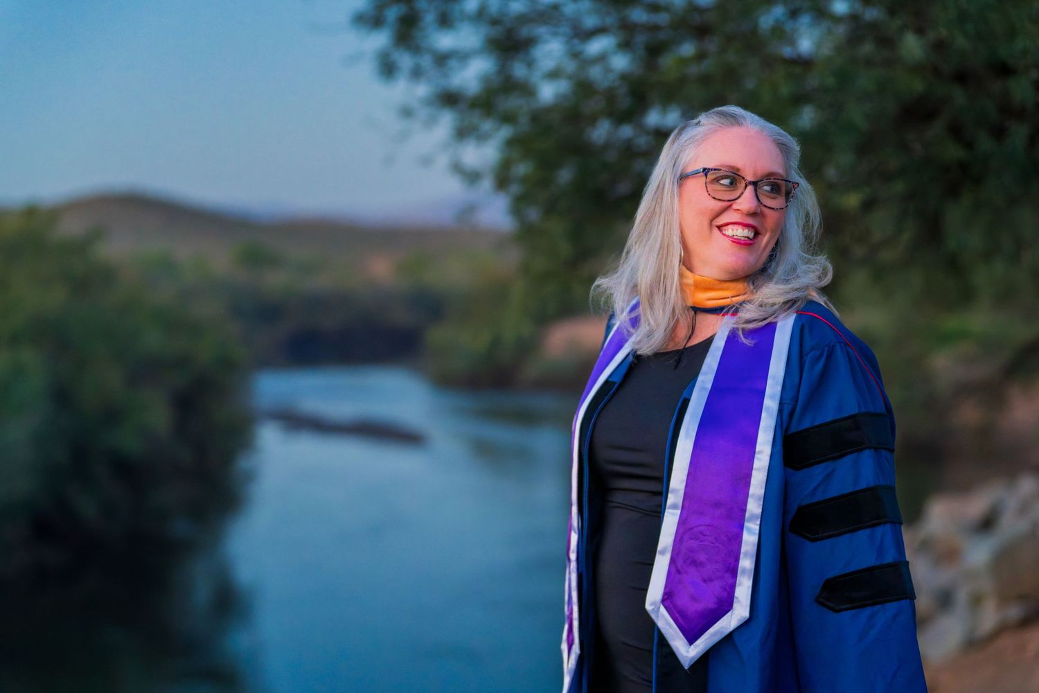 Graduate in academic regalia poses near scenic lake during golden hour.