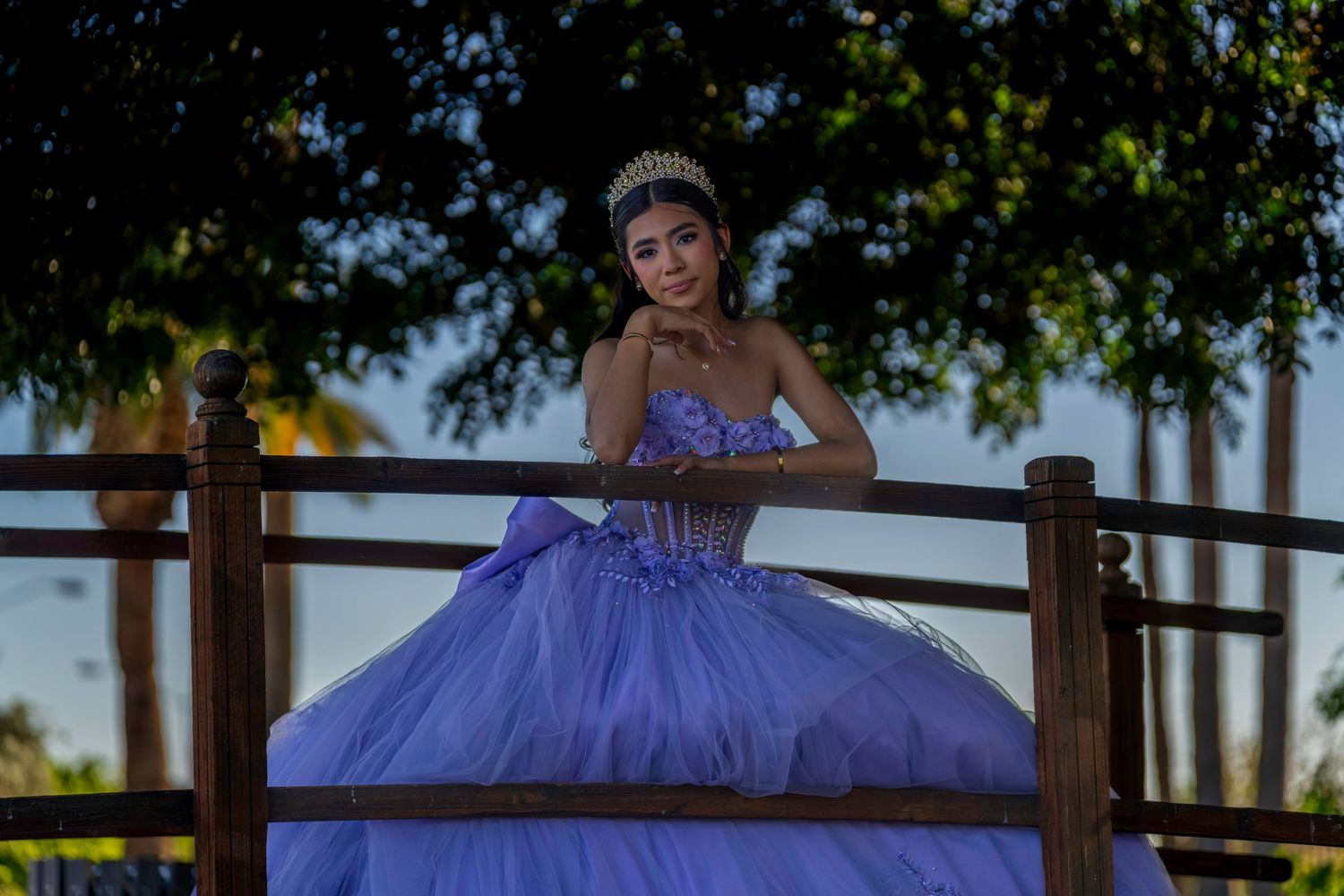 Elegant quinceañera girl in purple ball gown and tiara poses gracefully on wooden bridge at dusk.