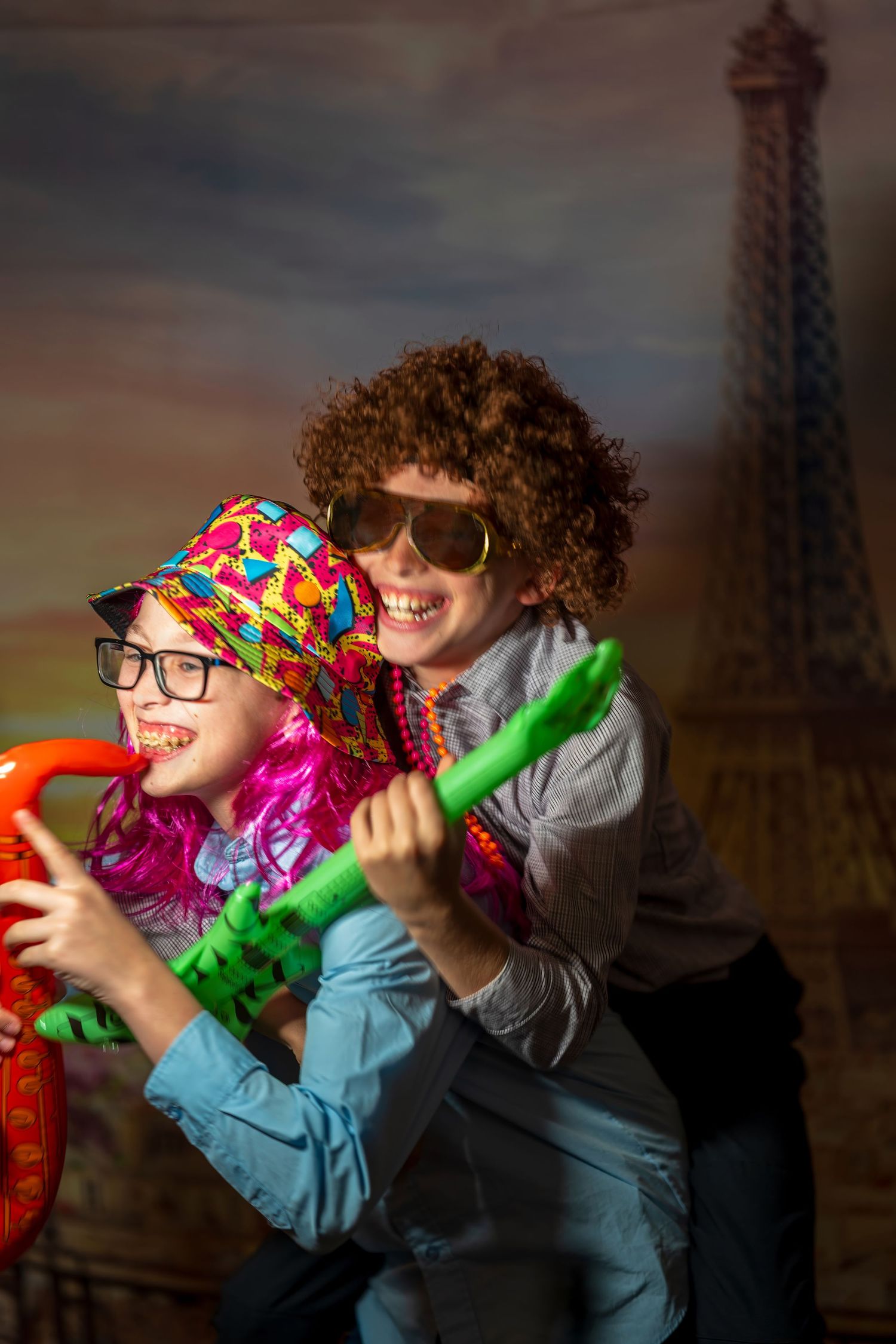 Two kids in colorful costumes and sunglasses pose playfully in front of an Eiffel Tower backdrop.