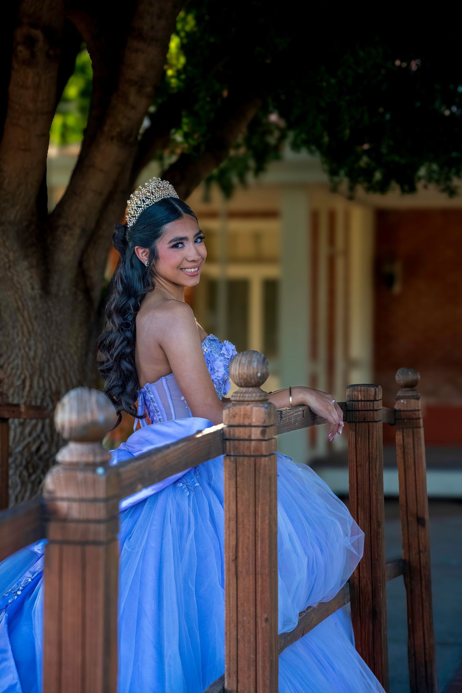 Young Latina woman in blue quinceañera gown and crown poses gracefully on wooden porch railing under a tree.