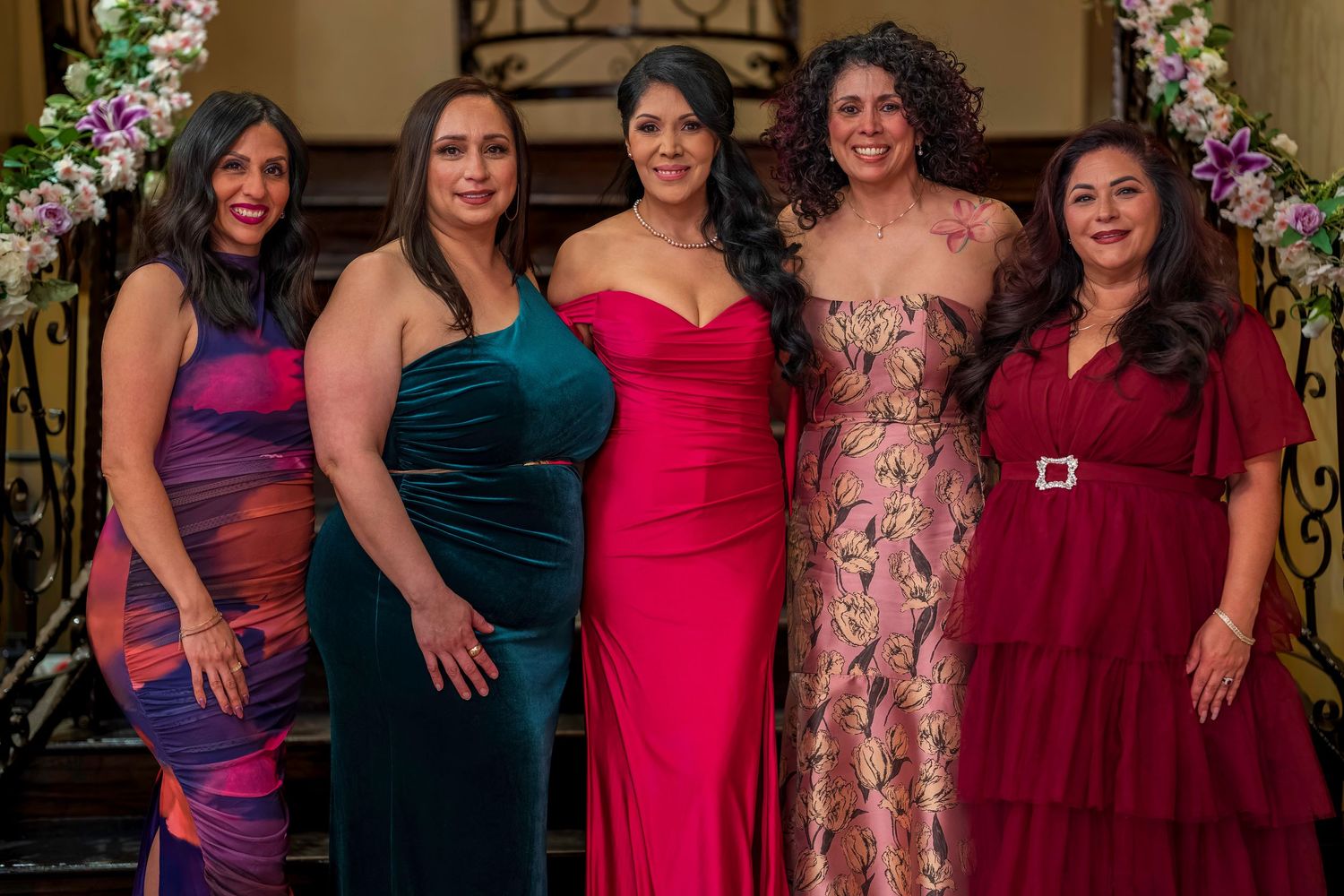 Five elegantly dressed women pose together in colorful formal gowns at a celebratory event near floral decor.