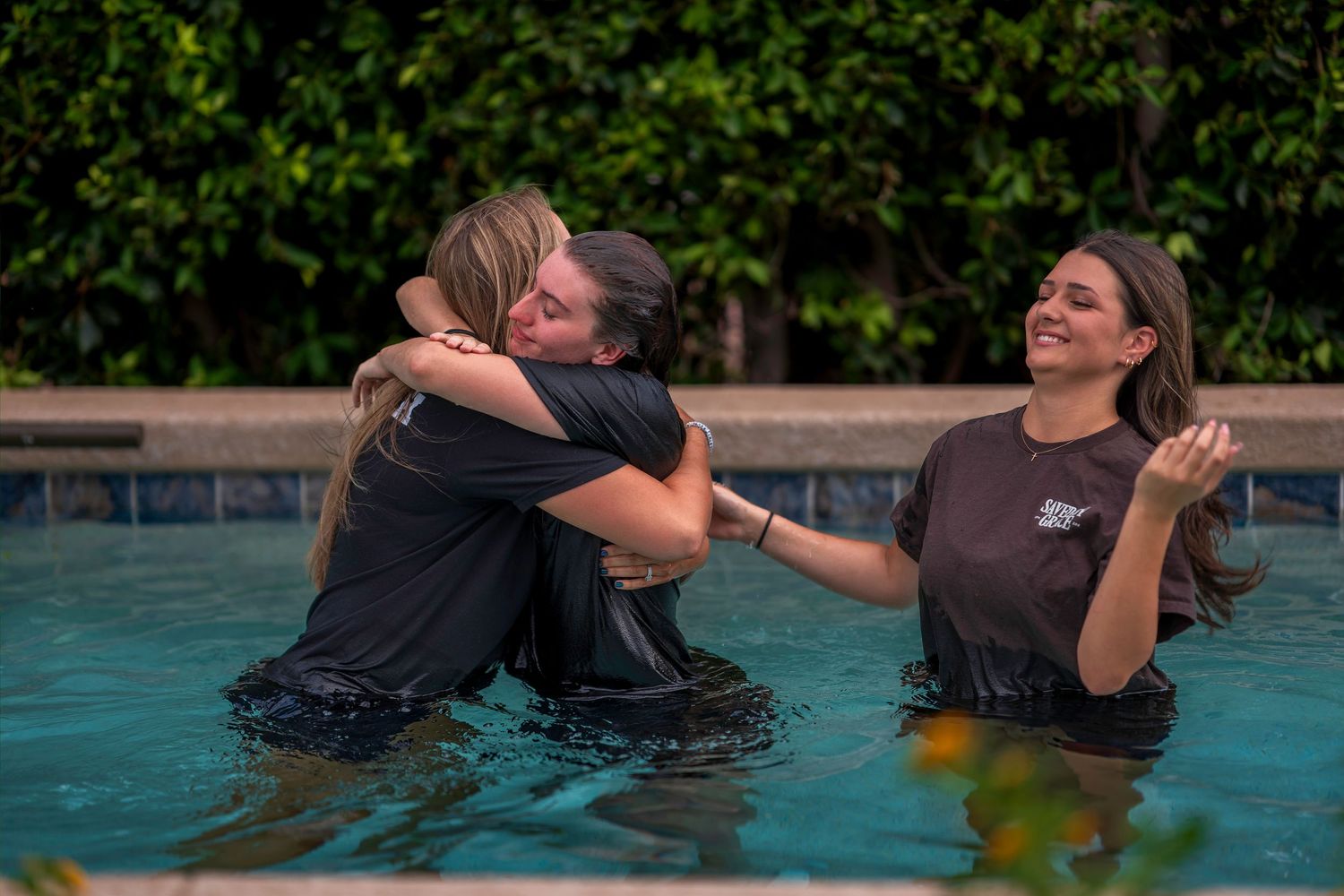 Three women sharing a joyful baptism moment in an outdoor pool, two hugging while a third smiles nearby.