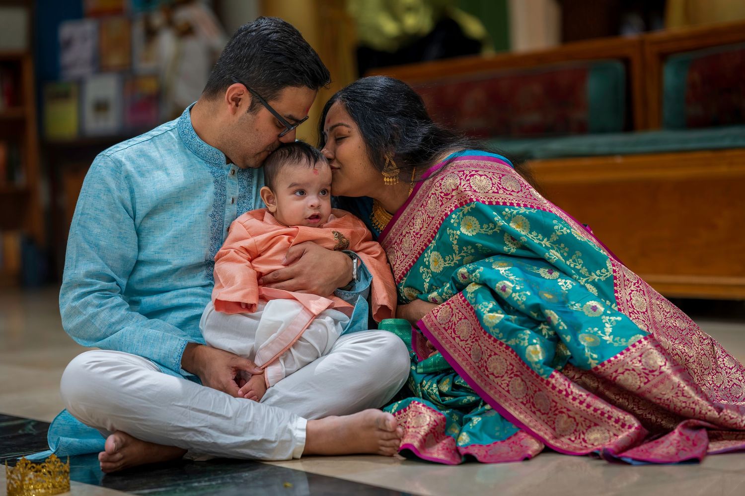 Indian couple in traditional attire sitting on floor, lovingly kissing their baby during a cultural ceremony.