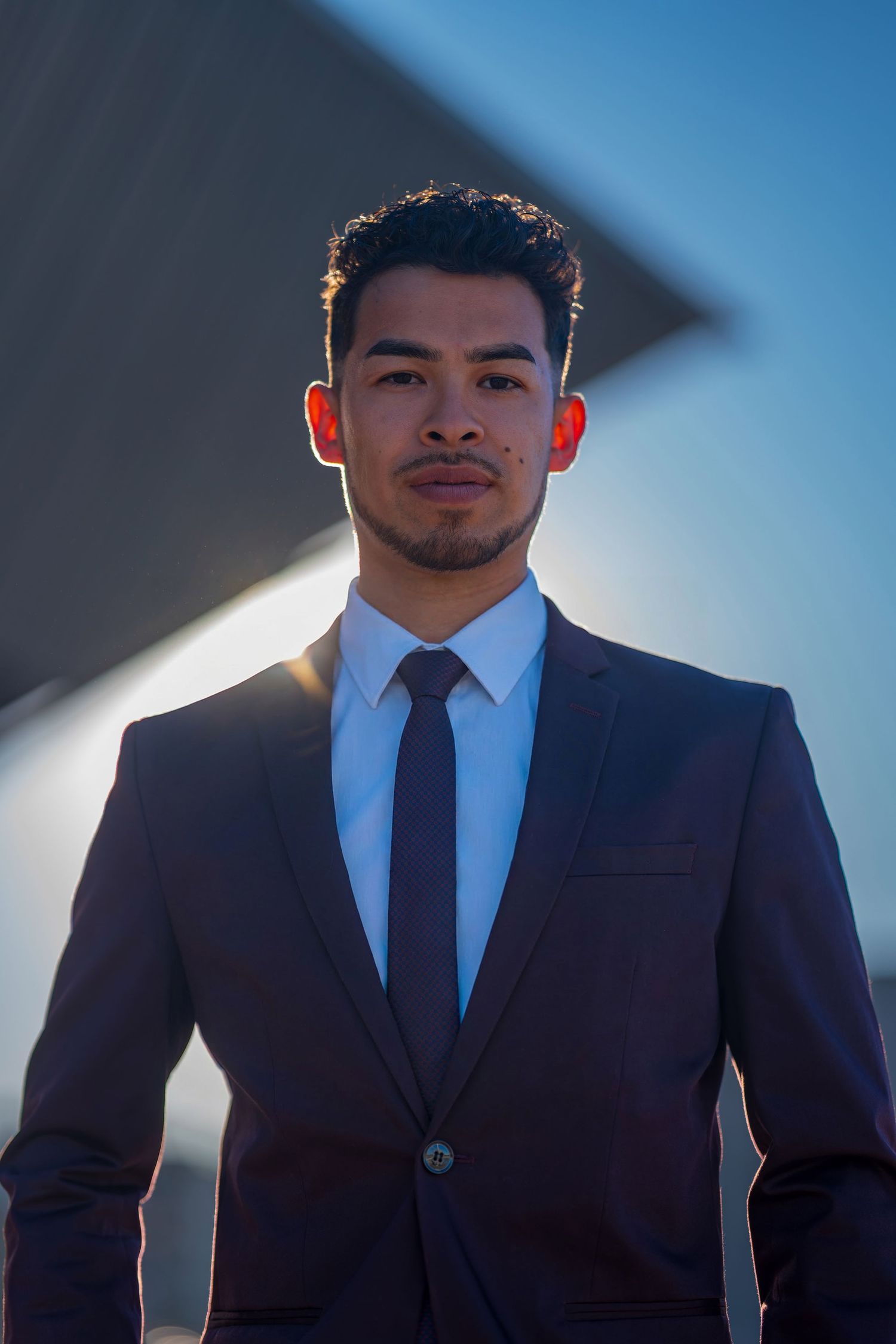 Professional headshot of person in dark suit and tie against blue sky backdrop.
