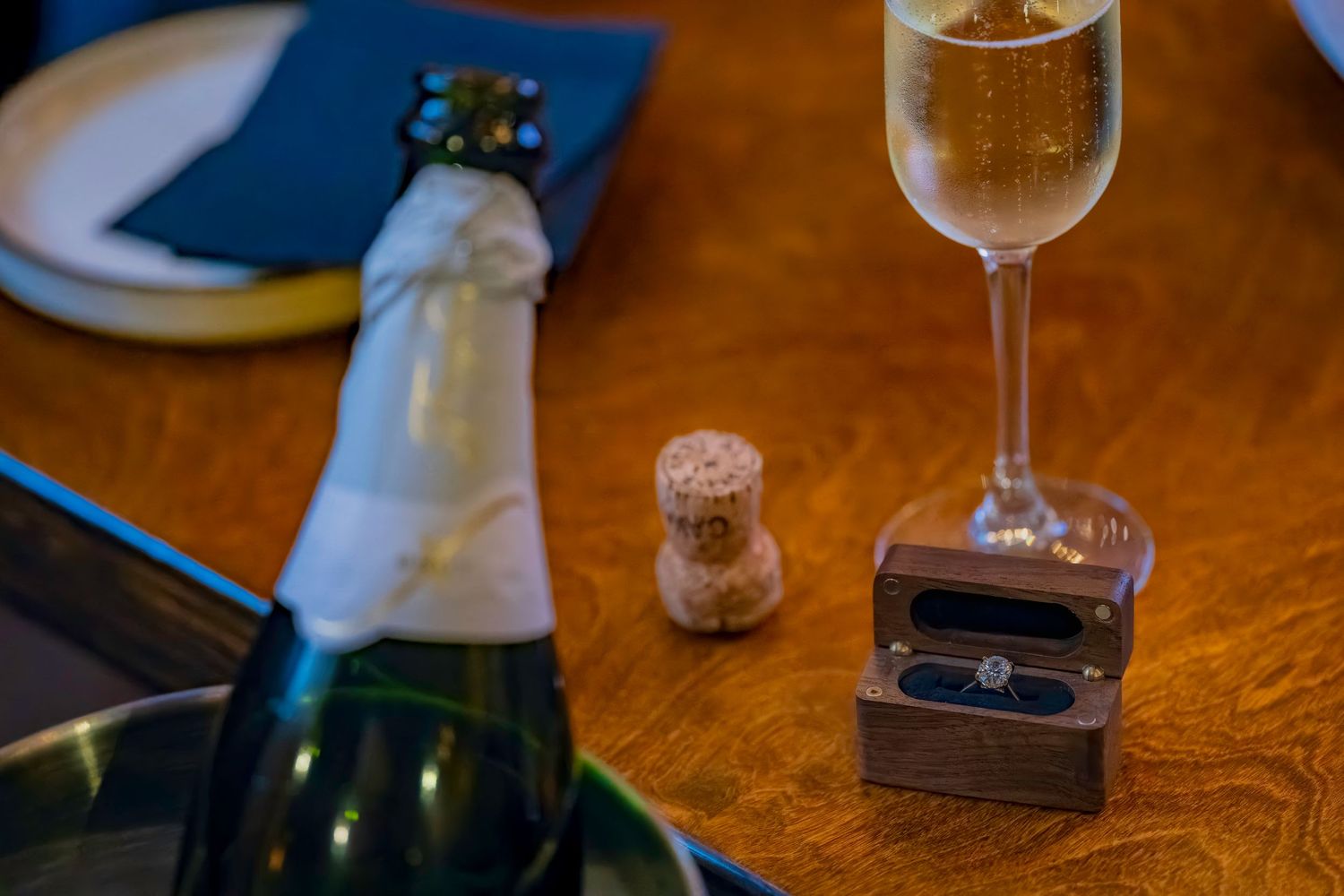 A champagne glass and ring box on a wooden table with wine bottle in view.