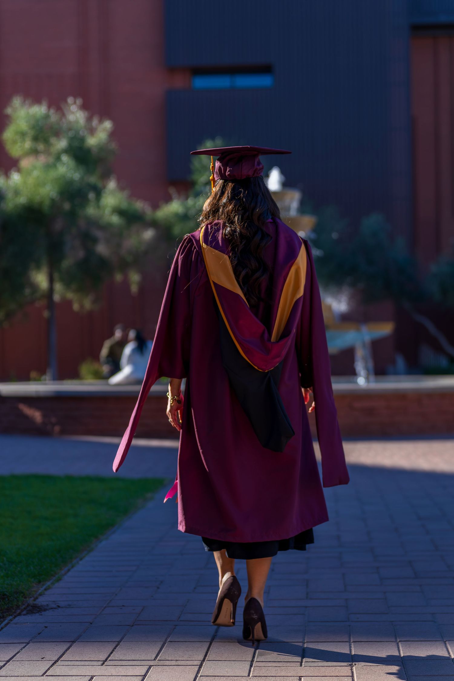 Graduate in maroon regalia walking away on campus path at dusk.