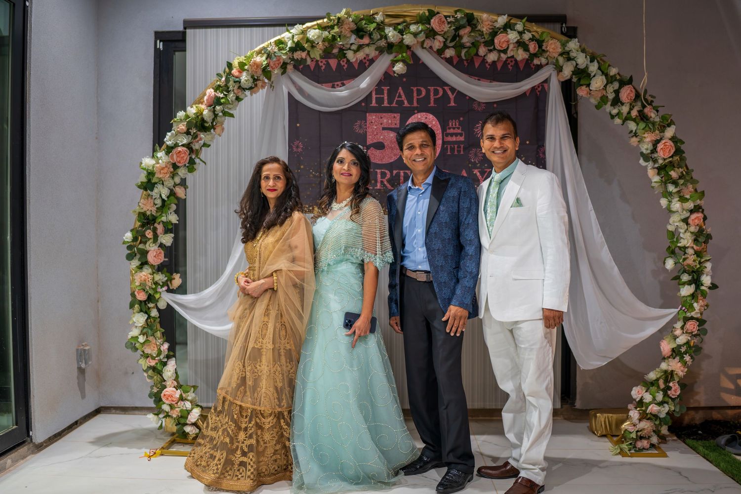 A group poses under a decorative floral arch with 'HAPPY' text during a celebration.