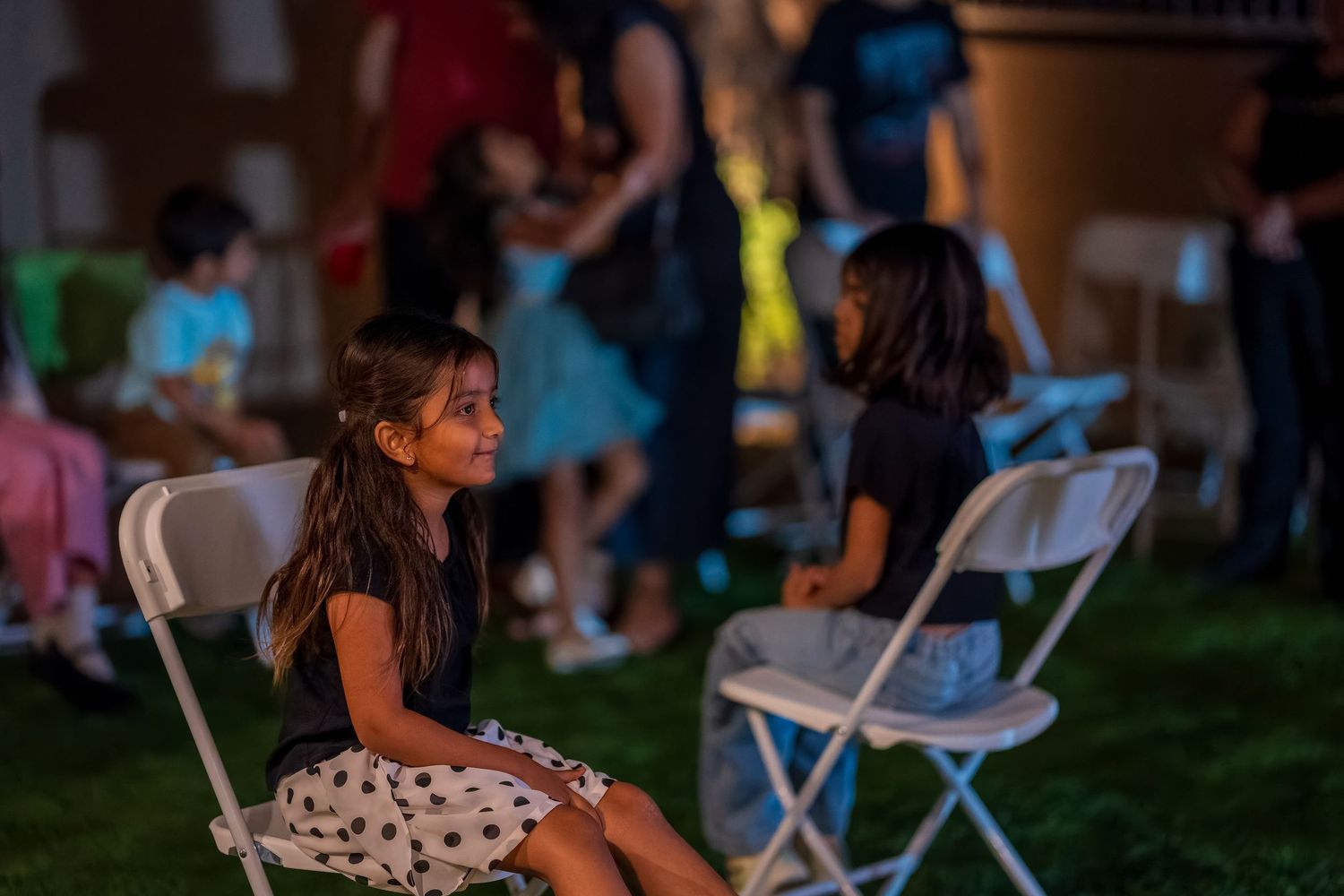 Child in polka dot dress sits on folding chair at dusk gathering.