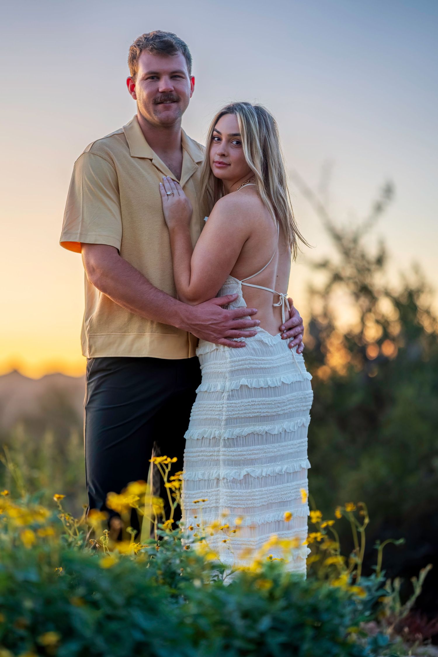 Couple embracing at golden hour sunset in a field of yellow wildflowers, woman in white dress, man in tan shirt.