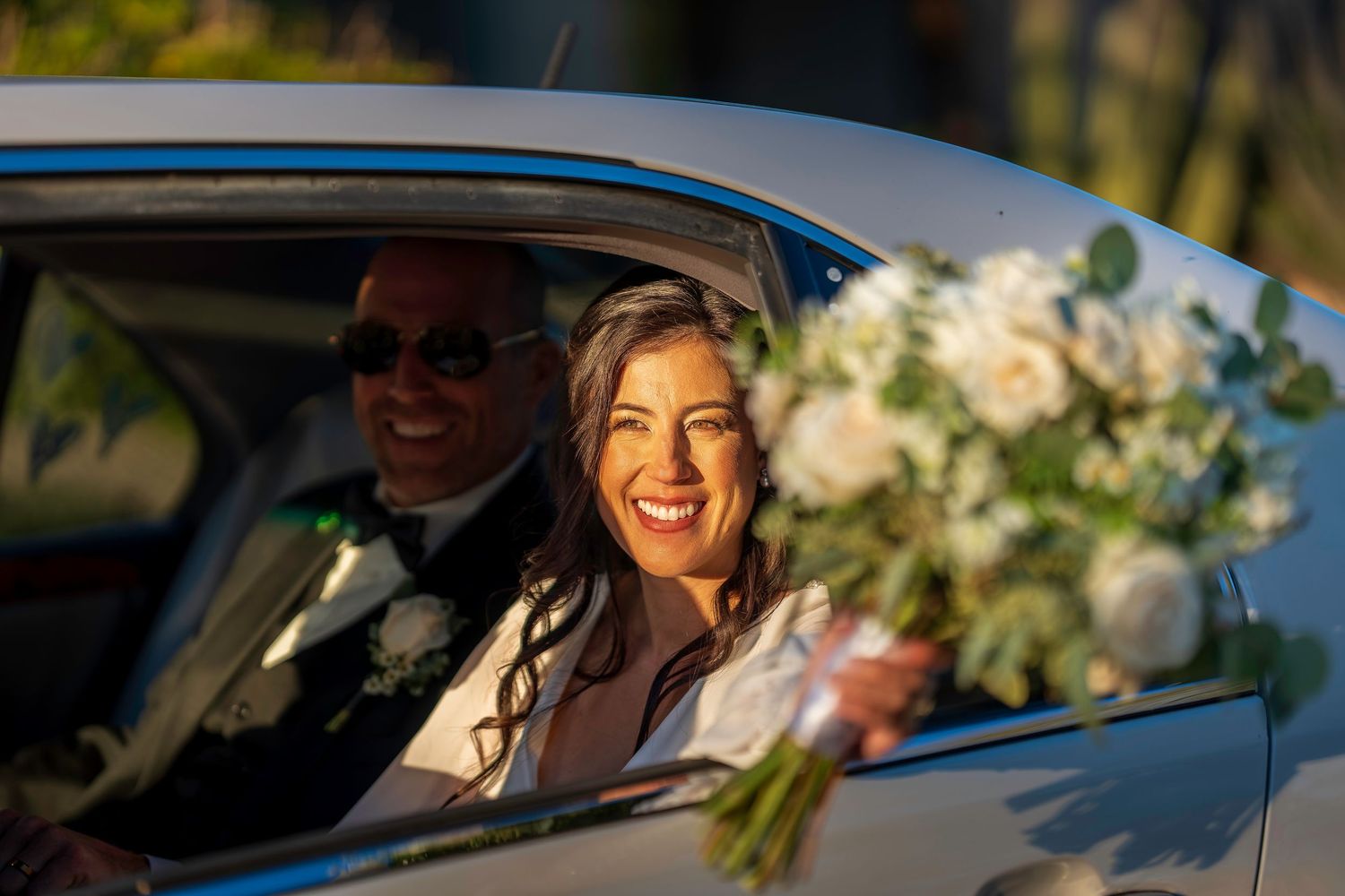 Radiant bride holding white floral bouquet smiles from car window with groom beside her.