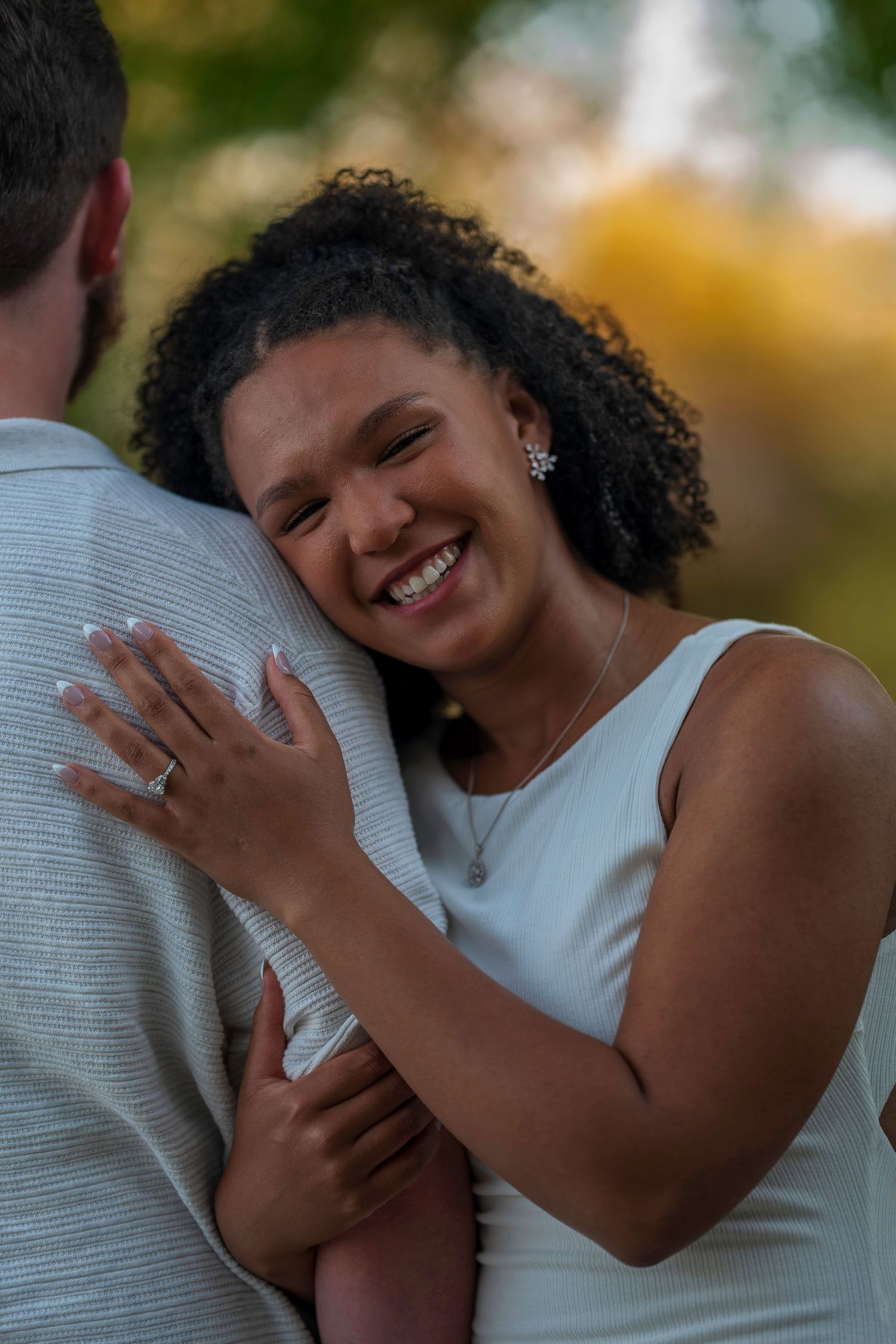Joyful woman in white dress smiling broadly while hugging her partner, showing engagement ring outdoors.