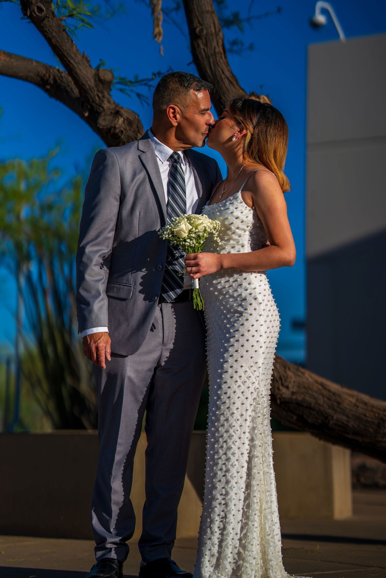 Bride and groom kissing outdoors, she holds white bouquet in fitted gown, he wears gray suit.